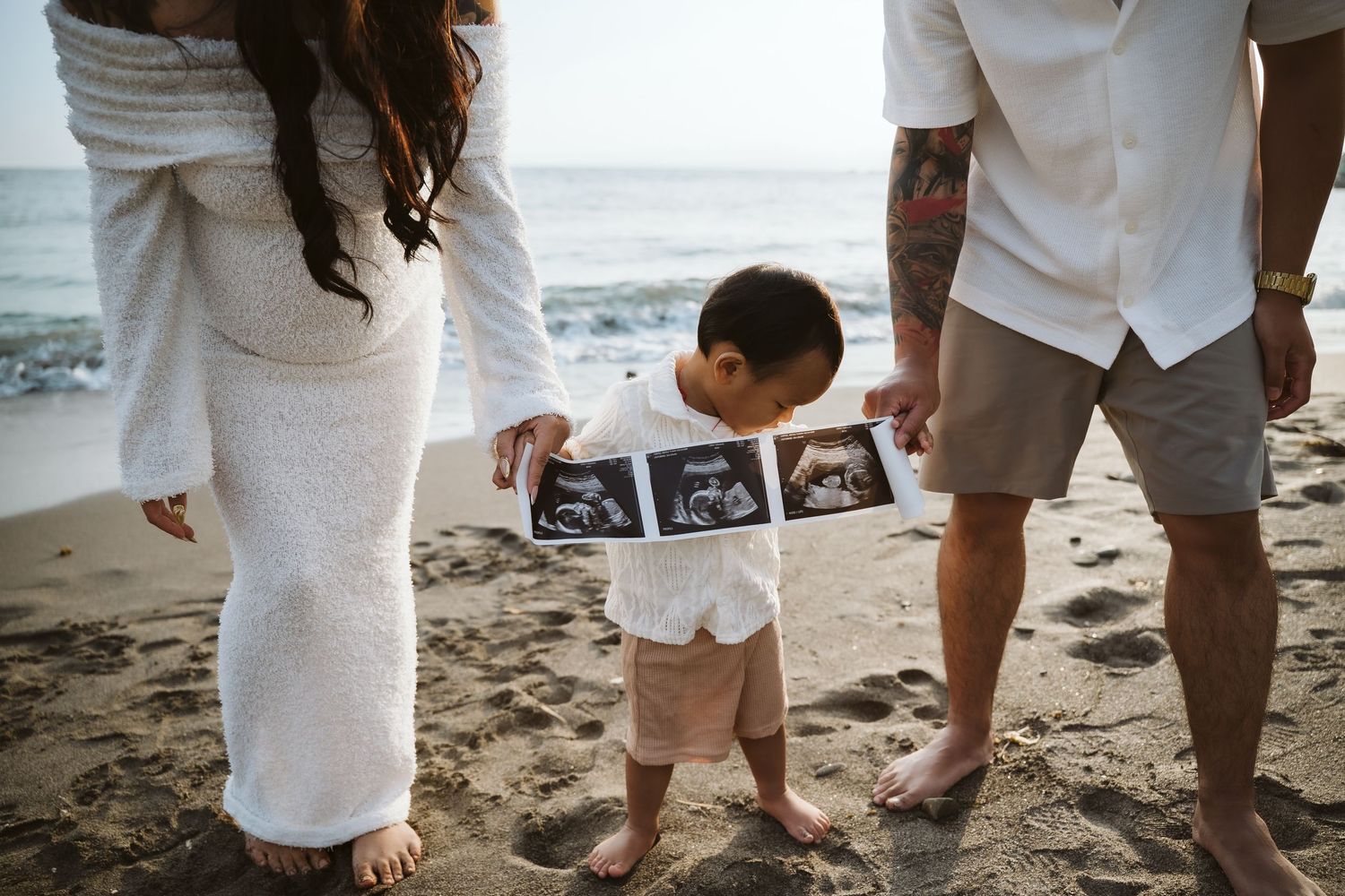 A toddler holds ultrasound photos on a beach while standing between two people in white outfits at sunset.