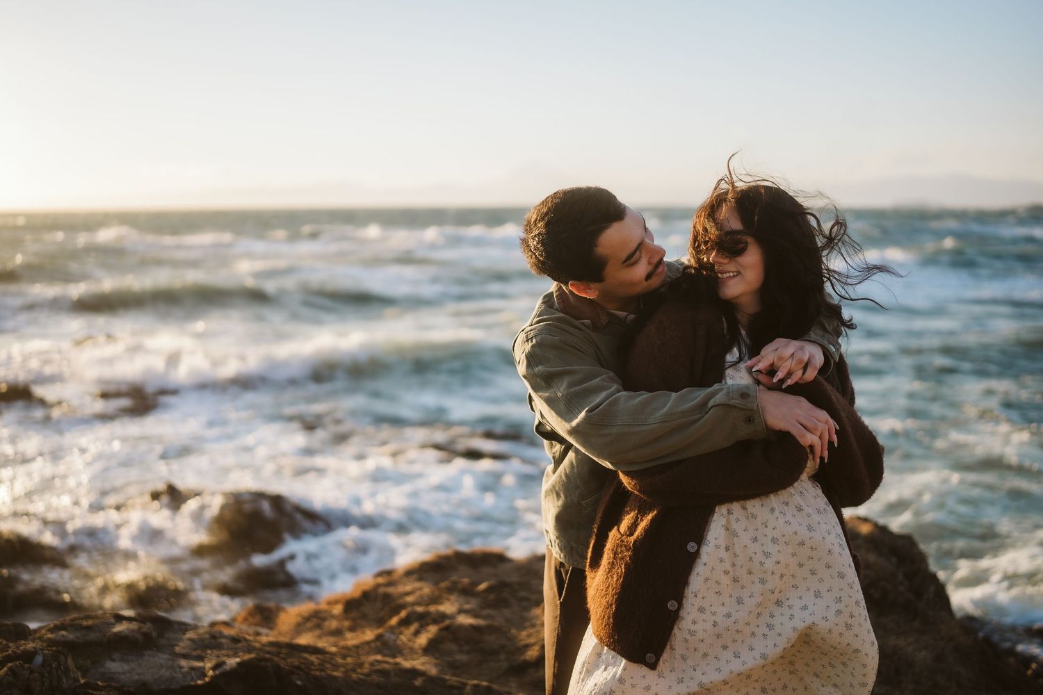 Intimate beachside moment captured against a backdrop of crashing waves and rocky shoreline.