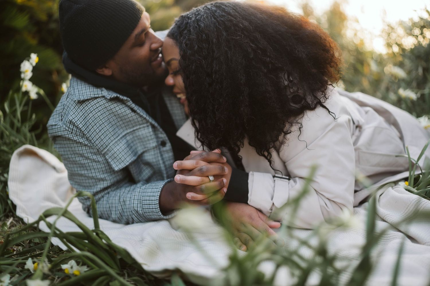A couple shares a tender moment while laughing together outdoors among white flowers in natural sunlight.