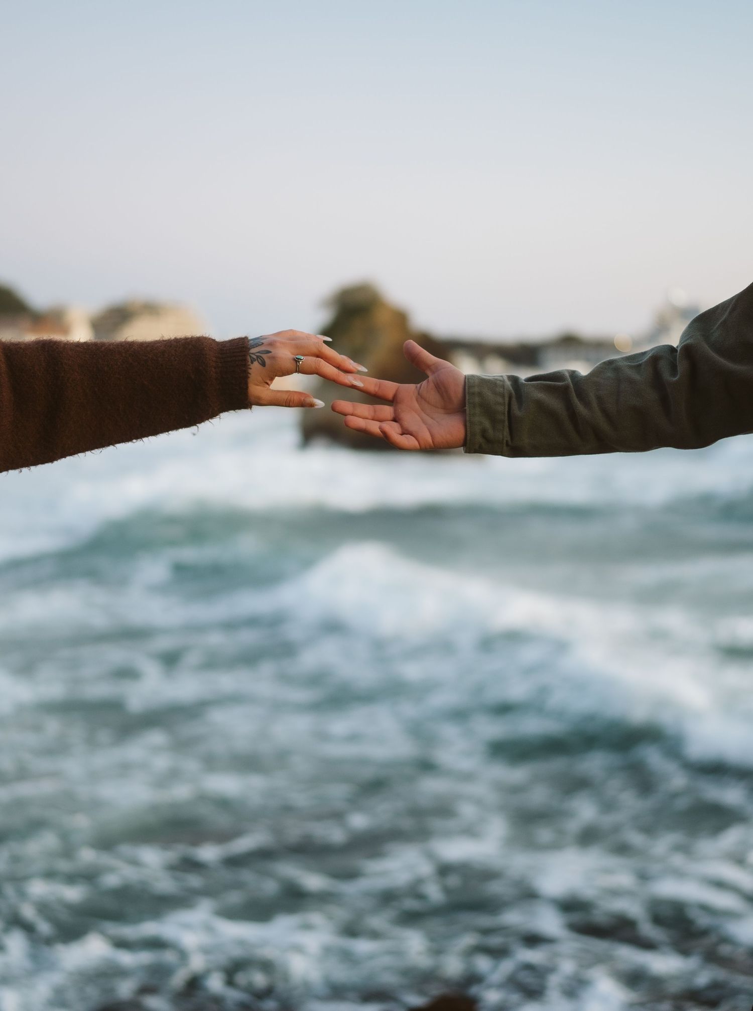 Outstretched hands reaching across ocean waves with rocky coastline in soft focus background.