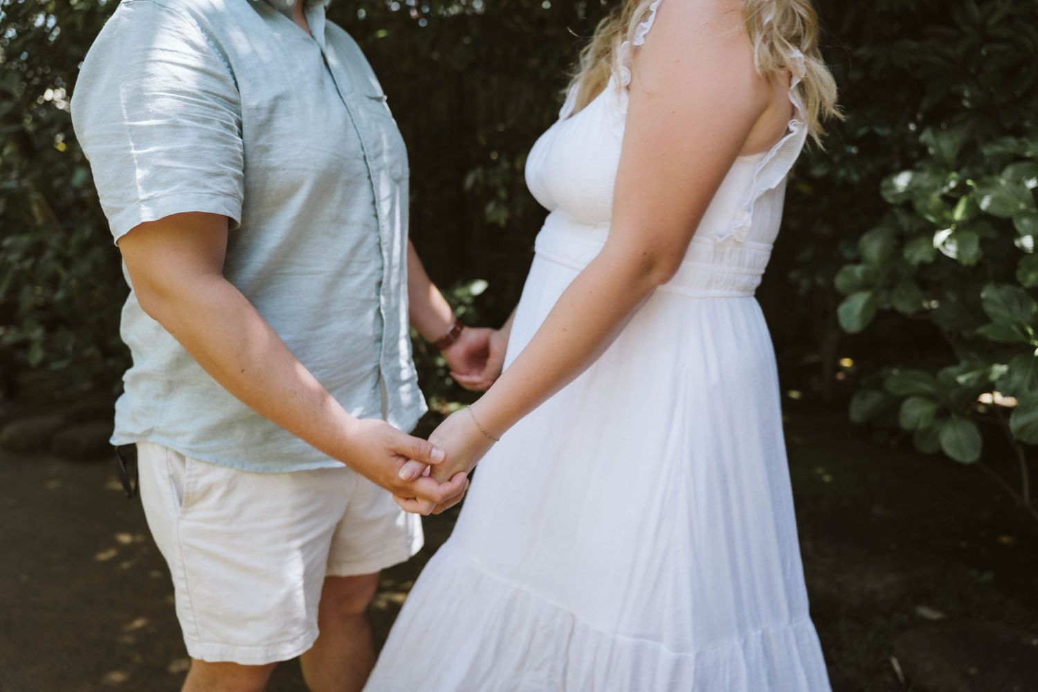 A couple in white attire holds hands during an intimate outdoor wedding ceremony in a garden setting.