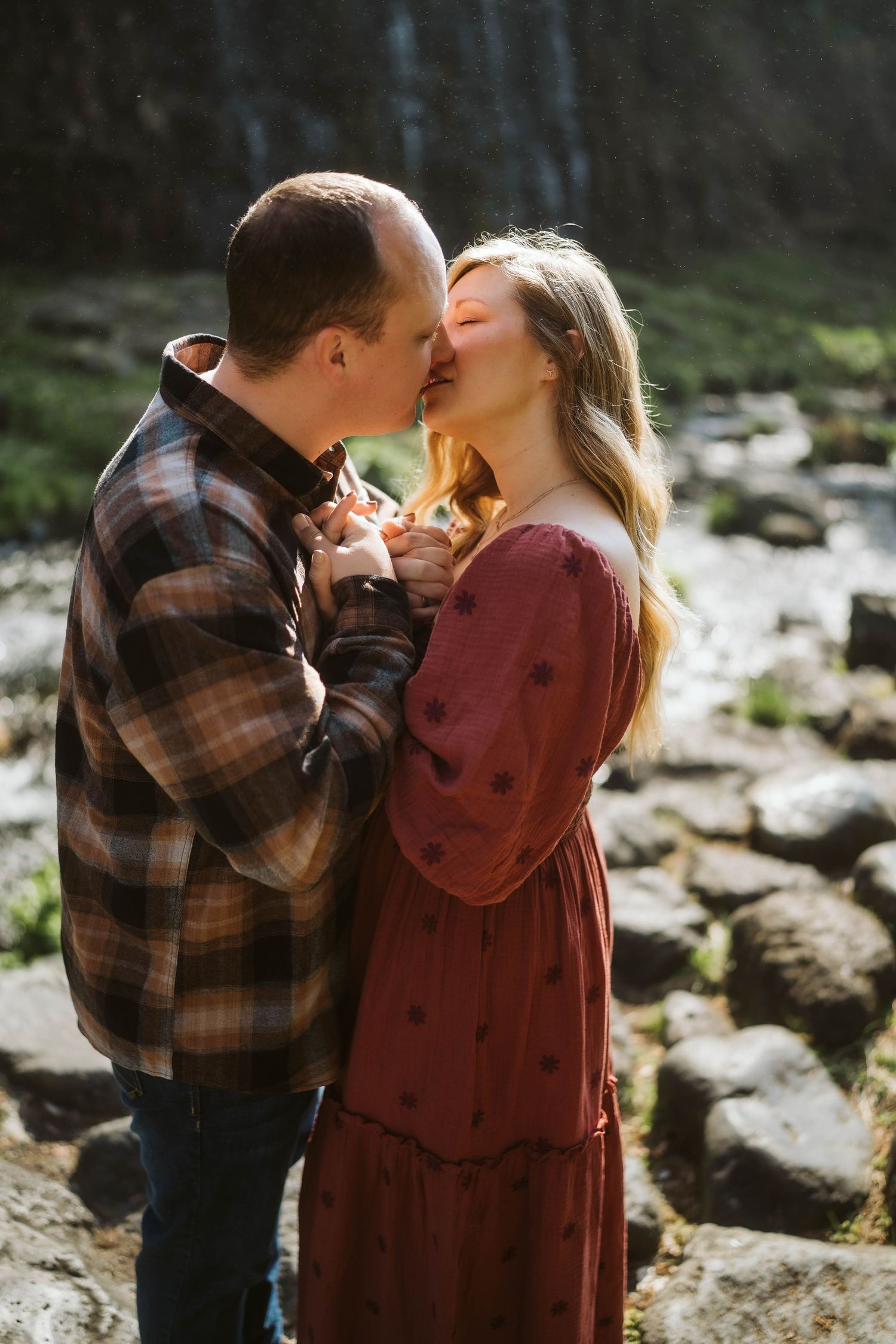 A couple in plaid and burgundy share intimate kisses against a rocky outdoor backdrop in natural lighting.