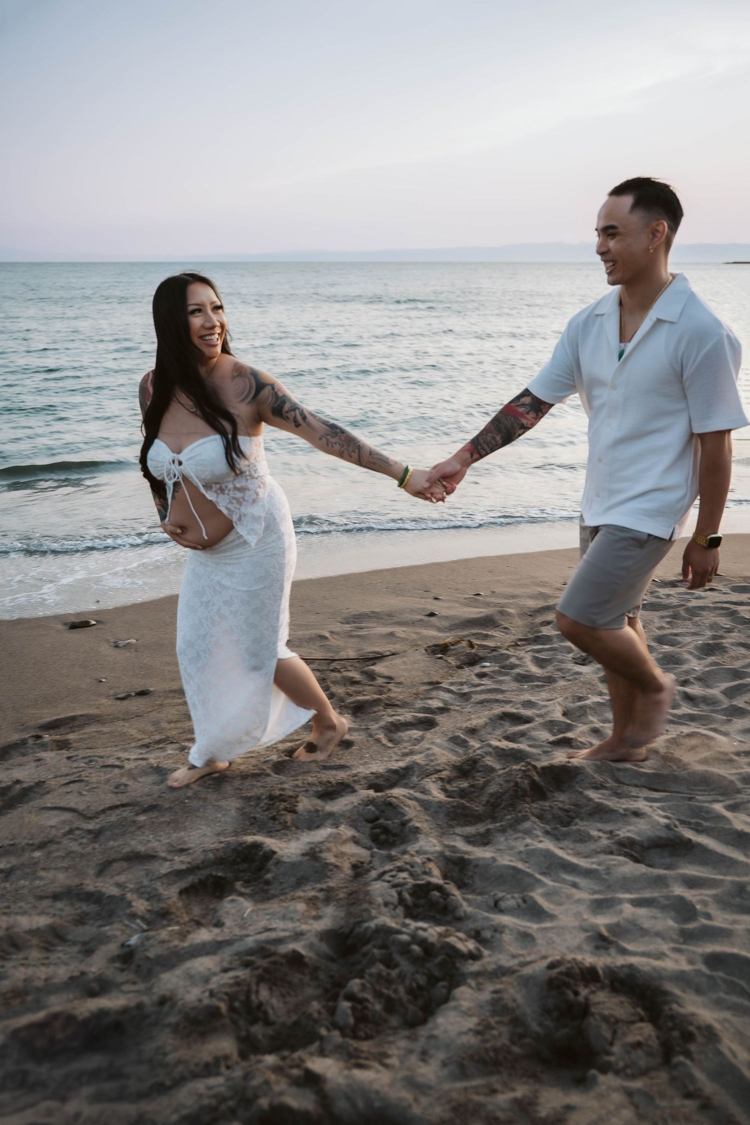 A couple in white clothing plays and laughs together on a beach with ocean waves in the background.