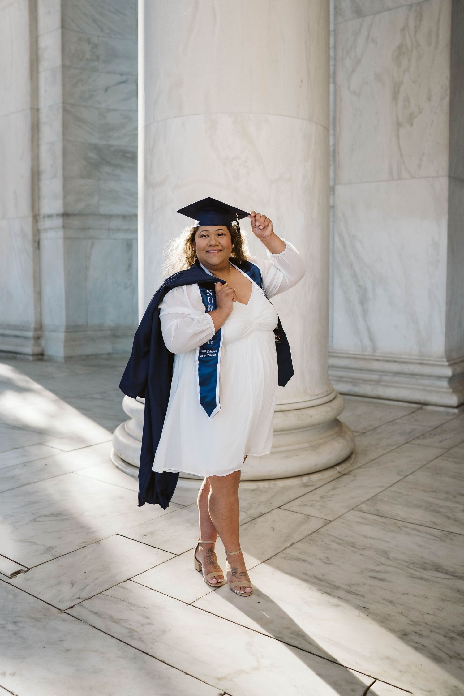 Graduate in cap and gown poses joyfully by marble columns with graduation attire and blue tie during commencement ceremony.