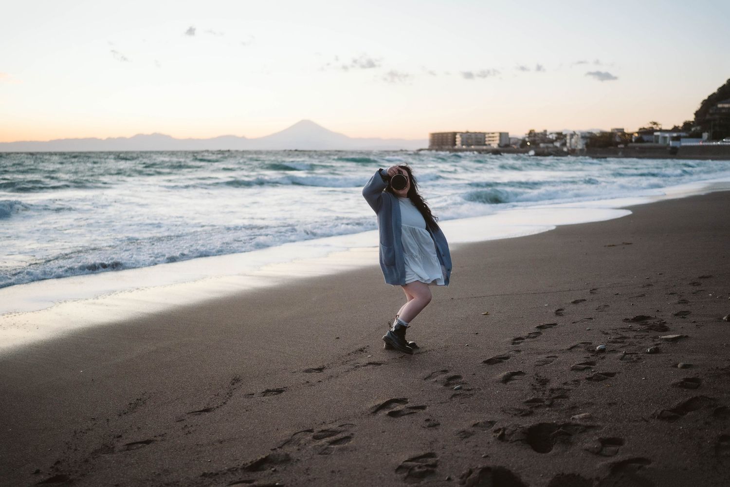 Person in denim jacket dances on a dark sandy beach at sunset with ocean waves and buildings in background.