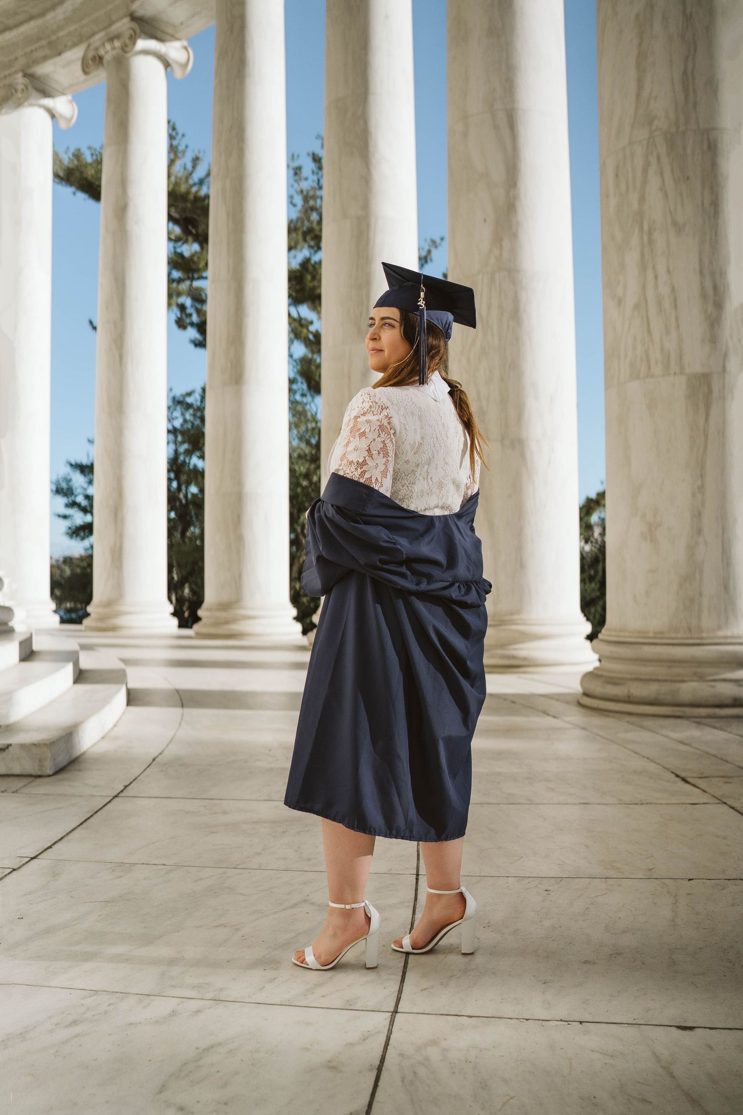 Graduate in blue cap and gown poses between classic white marble columns at university graduation ceremony.