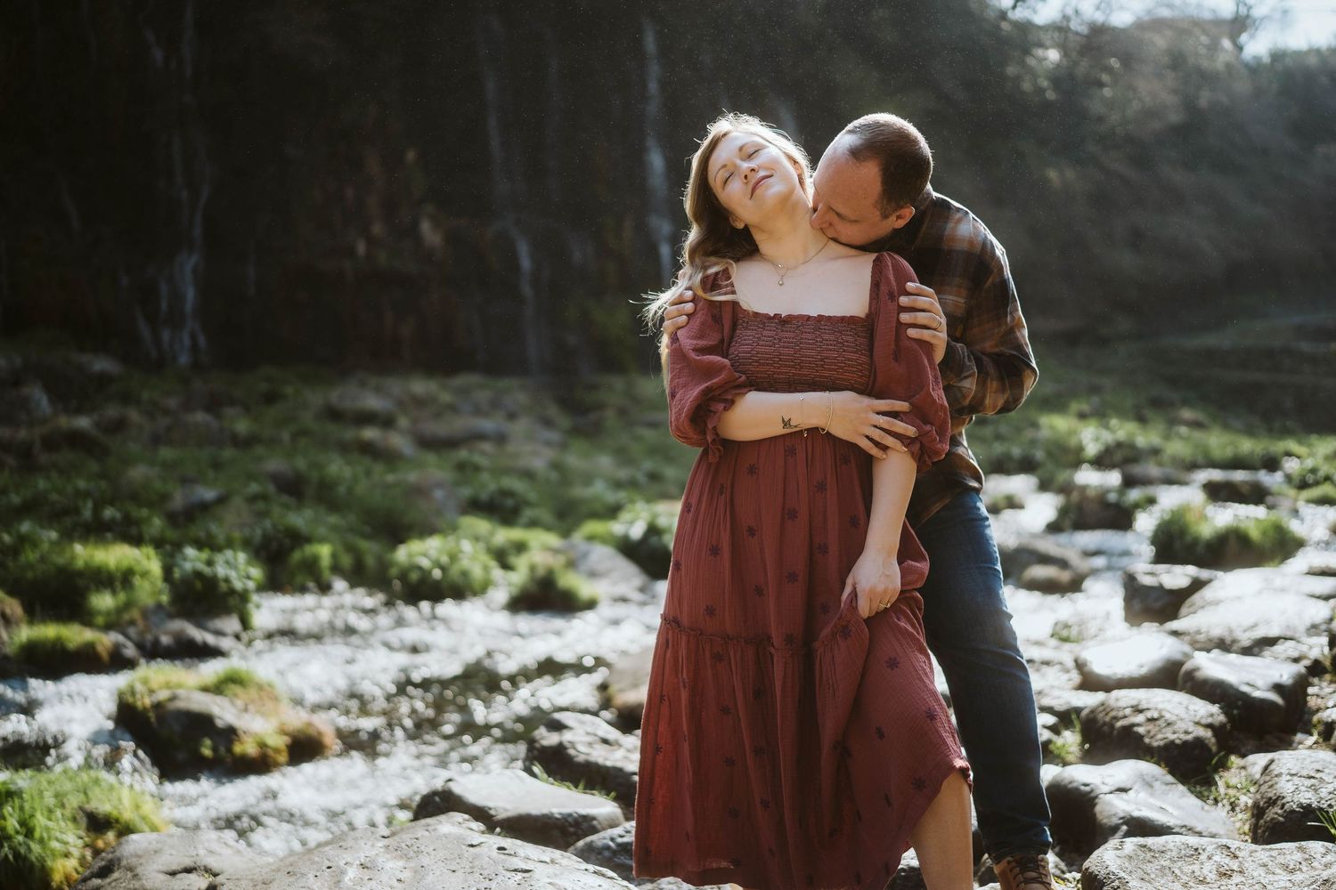 A couple in burgundy attire embraces while standing near a rocky stream in a misty forest setting.