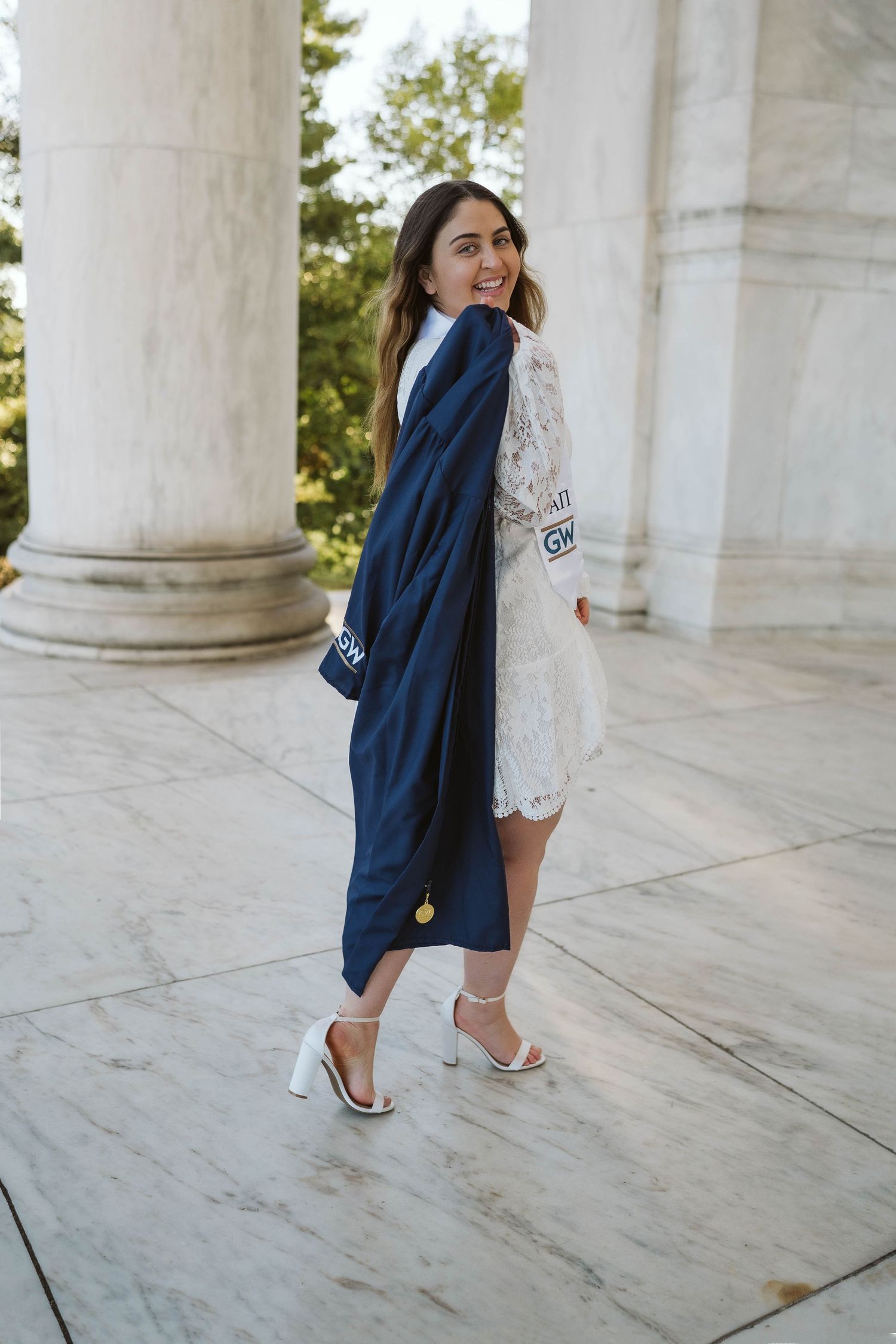 Graduate in white lace dress and navy blue graduation gown poses between marble columns at university ceremony.