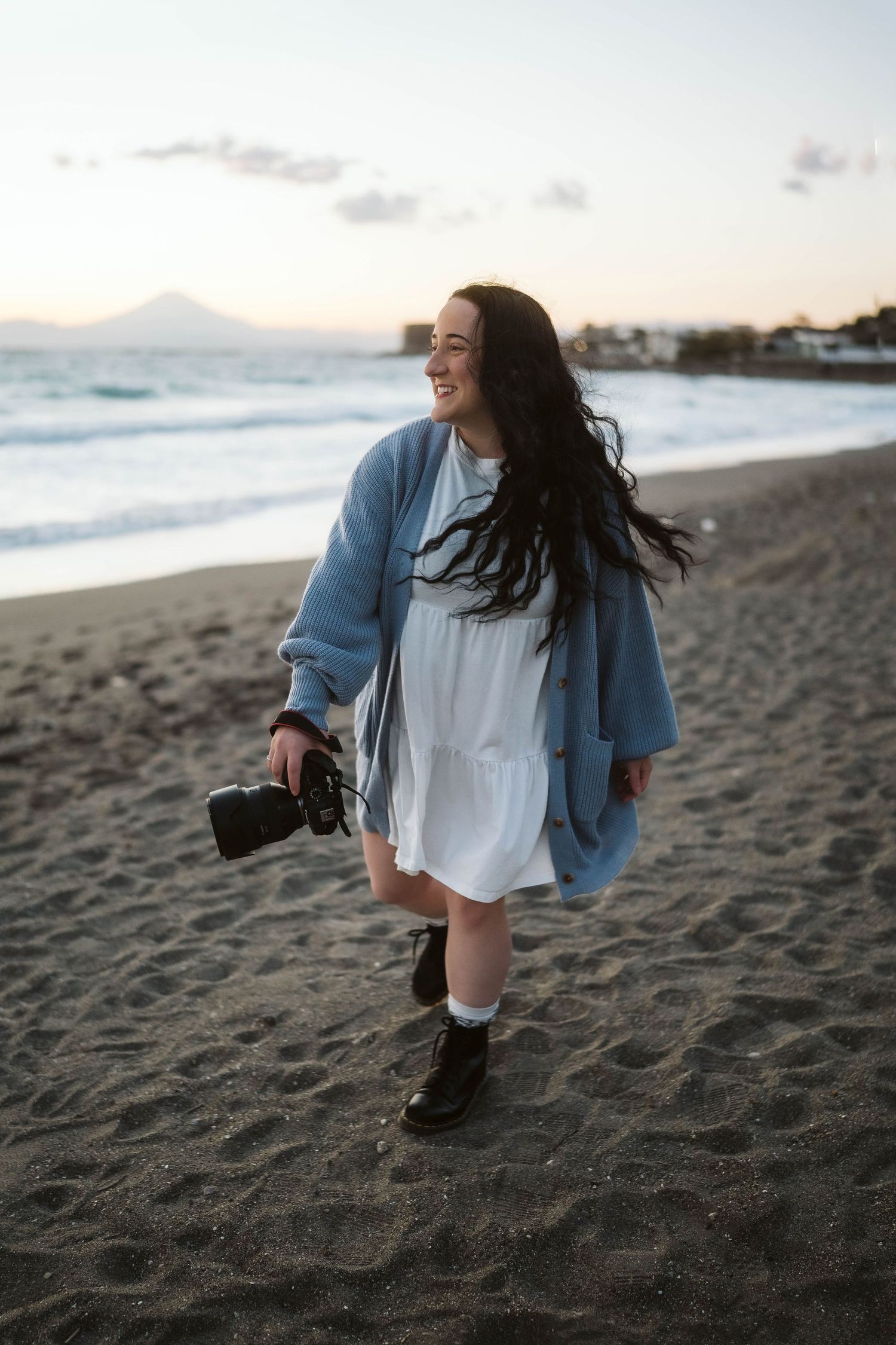 Person in a light dress and blue jacket walks joyfully on a beach at sunset holding a camera.