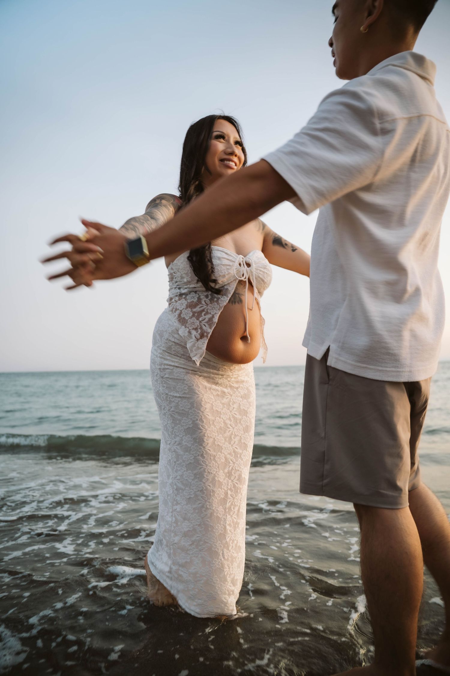 A couple dances playfully in the ocean waves, wearing white attire on a sunny beach.
