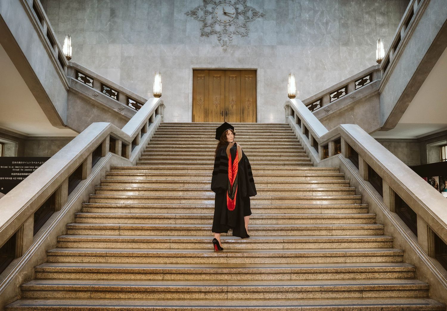 Wide marble staircase in a grand building with figure in graduation attire at the top.