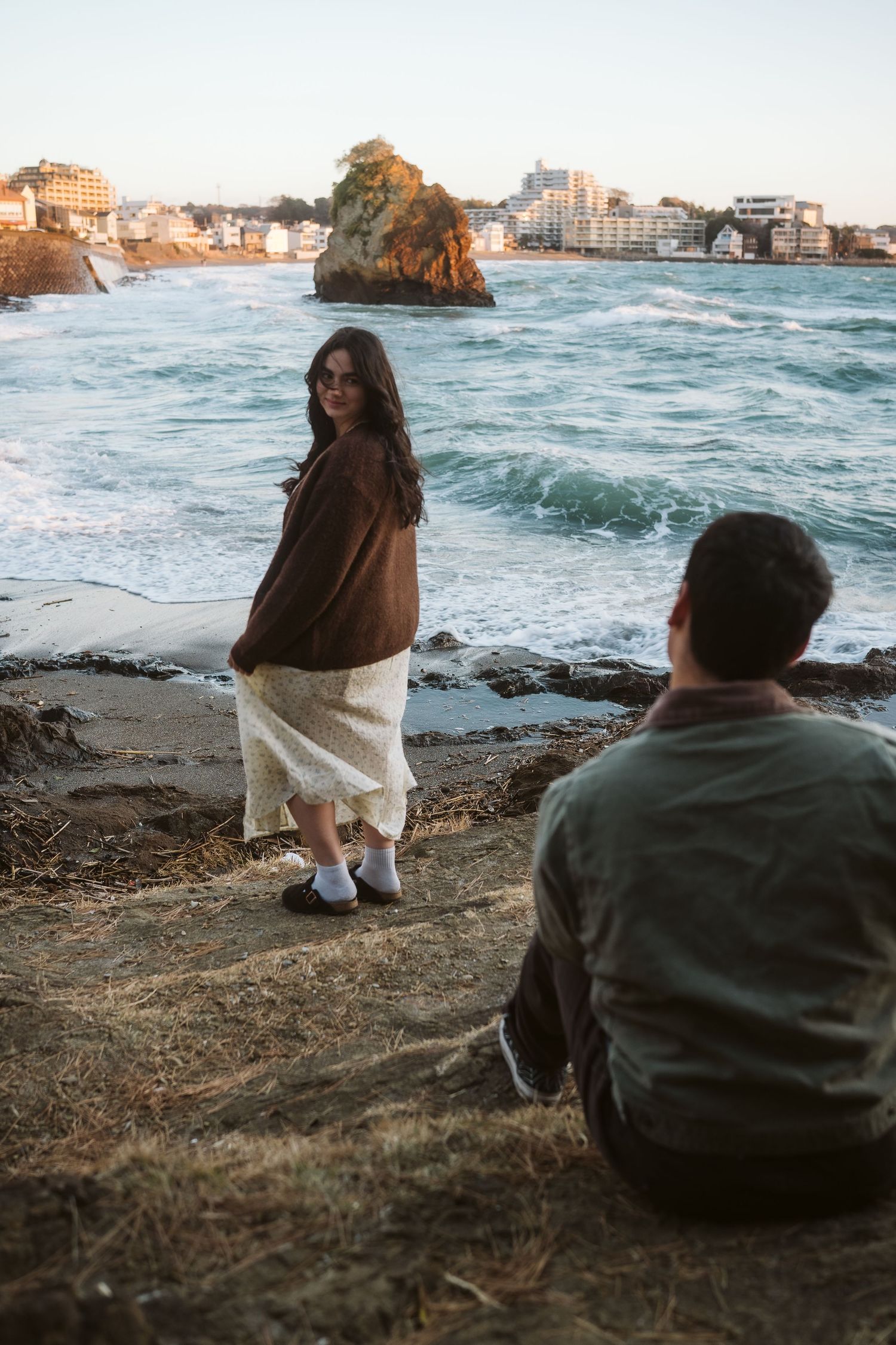 Couple relaxing on coastal rocks overlooking the ocean and cityscape at sunset.