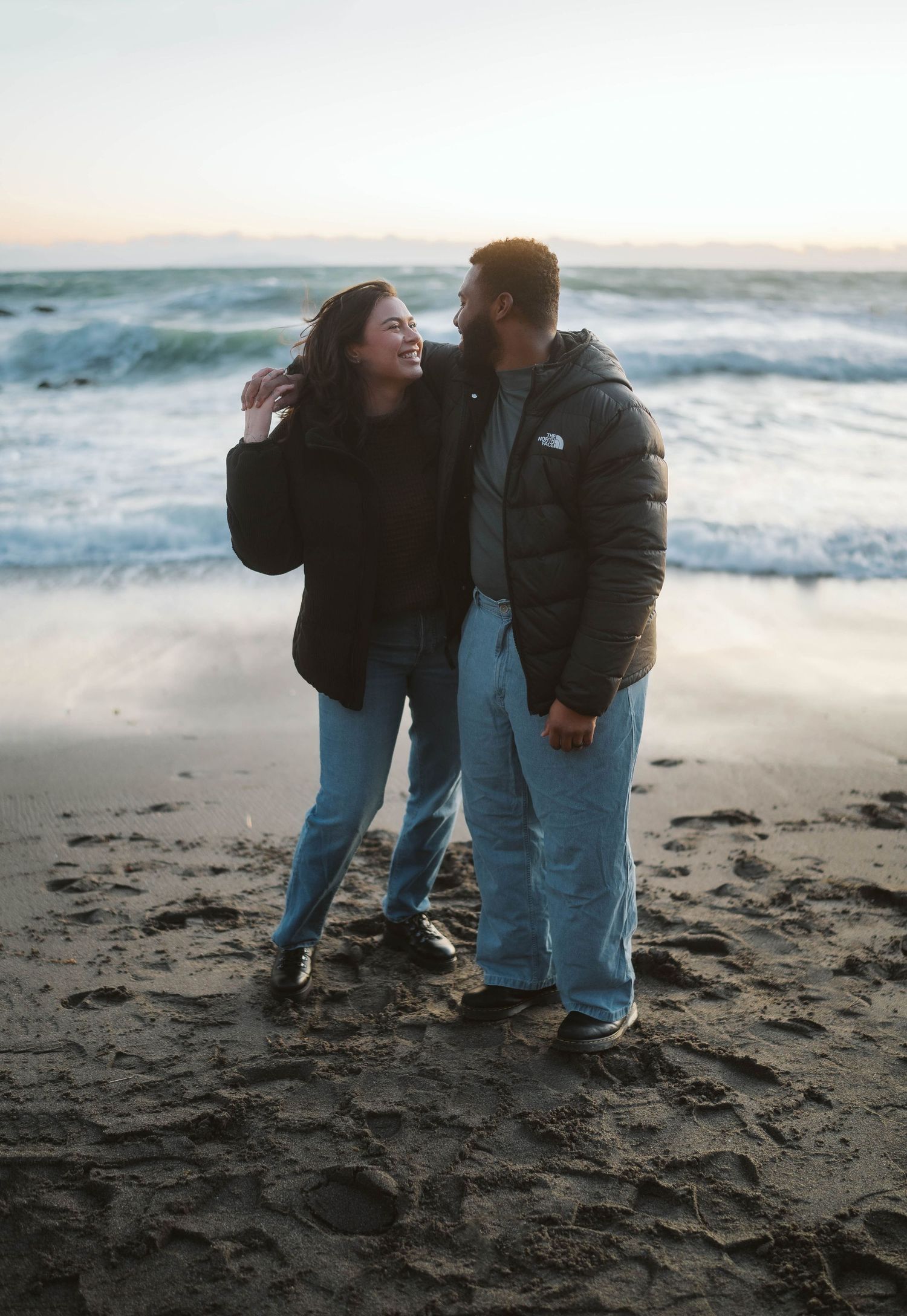 Two people walk together along a misty beach shoreline at sunset with waves crashing in the background.