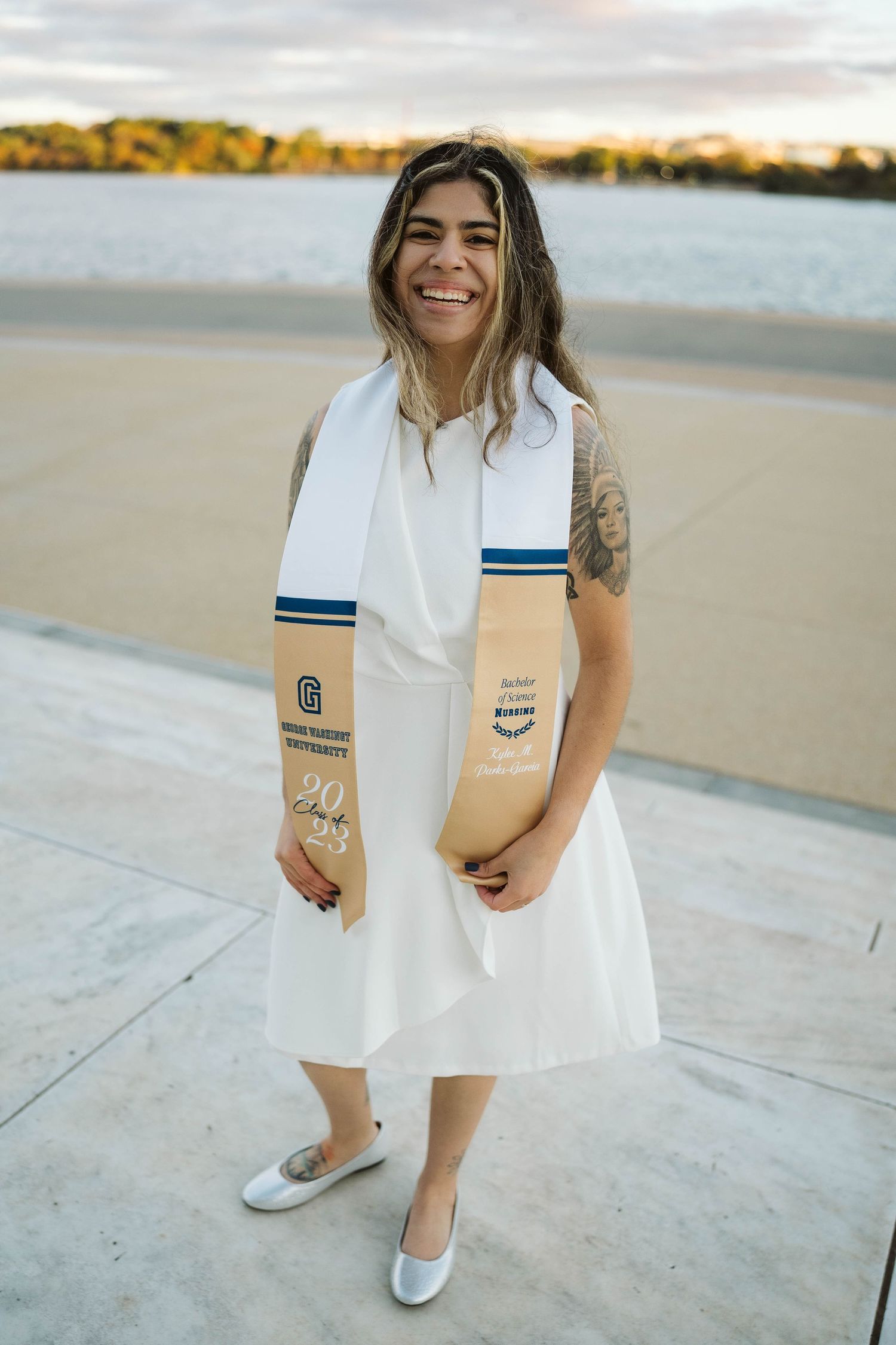 Graduate wearing white dress and ceremonial stole with blue stripes poses outdoors by waterfront at sunset.