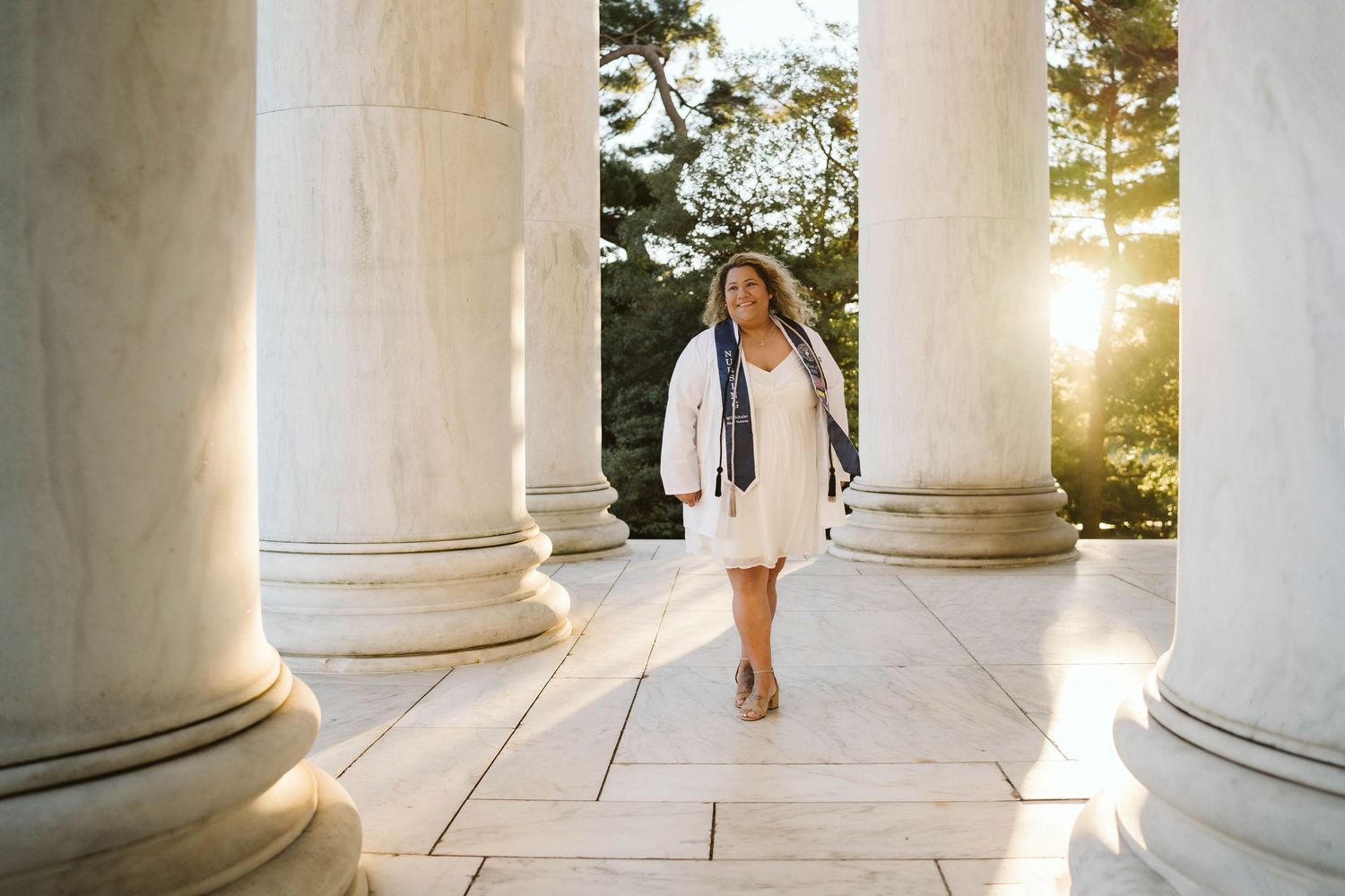 White dress graduation photo session between classical columns at sunset with lens flare effect.