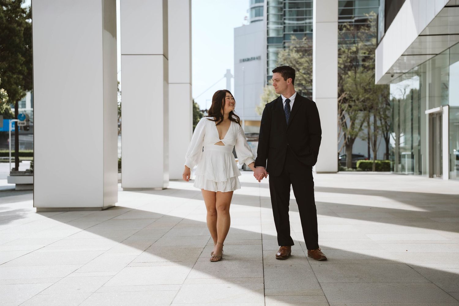 Two people in formal attire holding hands while walking together under a modern white architectural walkway.