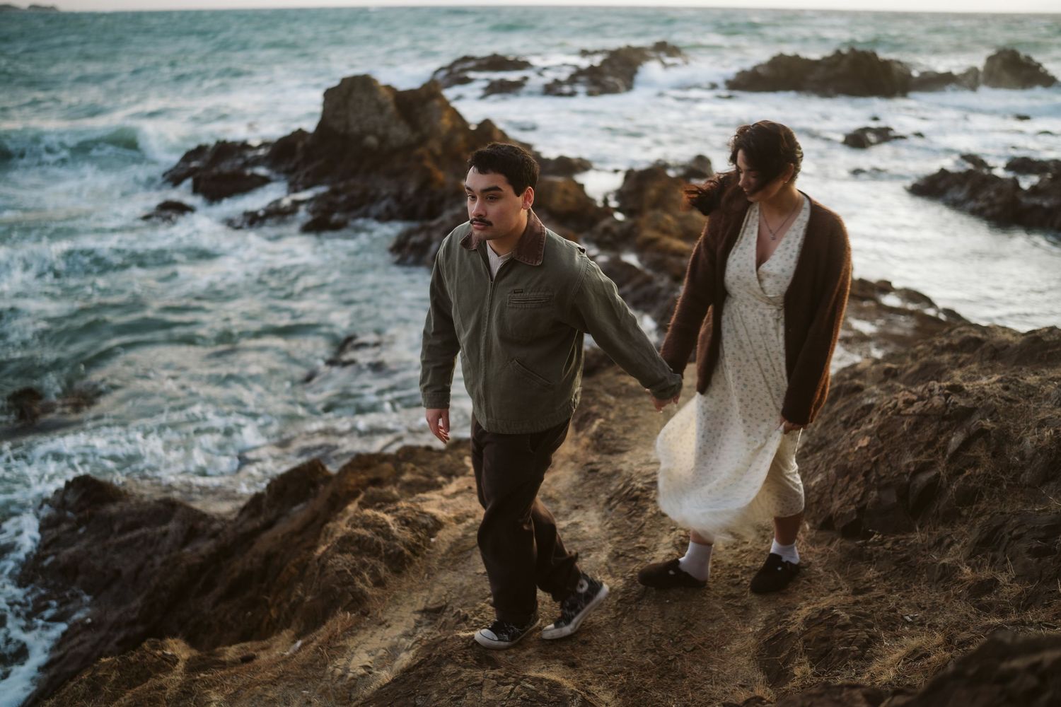 Scenic coastal engagement photos with dramatic waves crashing against rocky cliffs in the background.