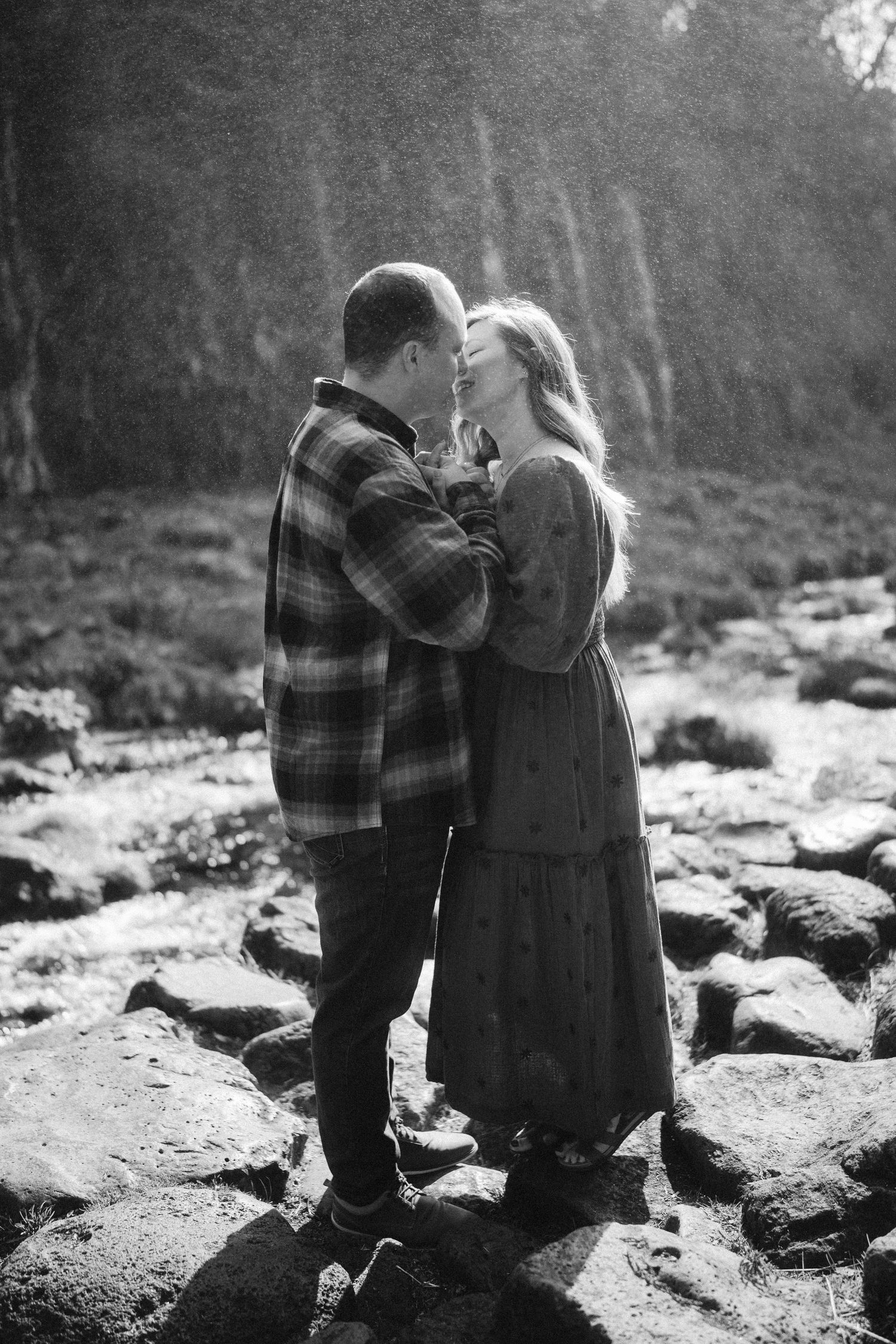 A romantic black and white photo of a couple sharing an intimate moment while standing on rocky terrain by a stream.