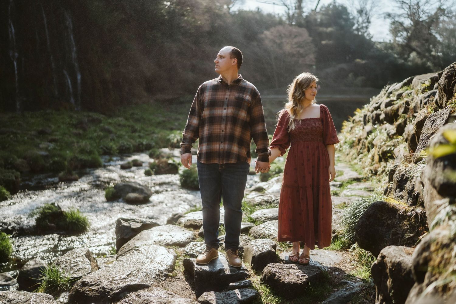 A couple holds hands while standing on rocky terrain near a stream in warm sunlight.