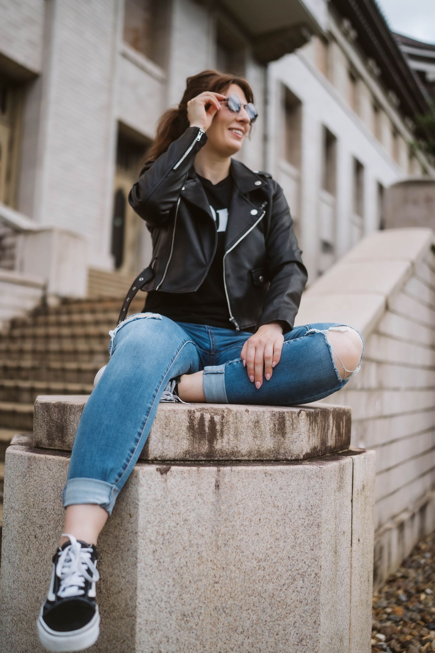 Person in black leather jacket and rolled up jeans sits casually on concrete steps wearing sneakers.