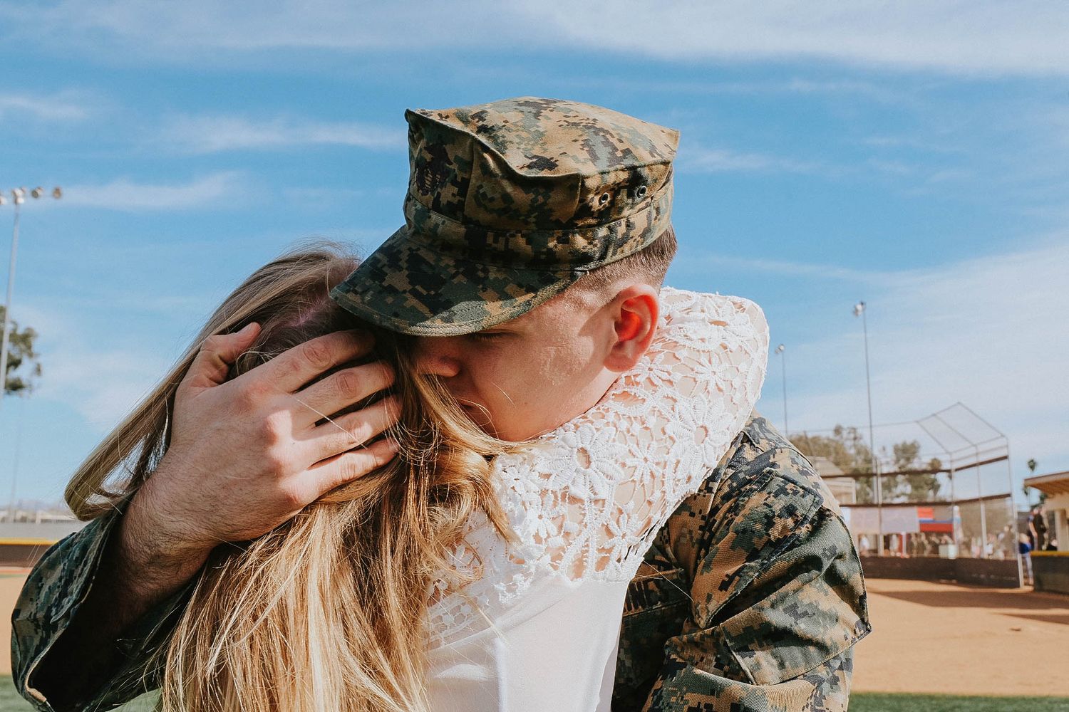 A person in military camouflage uniform and cap embraces another person wearing white lace outdoors on a sunny day.