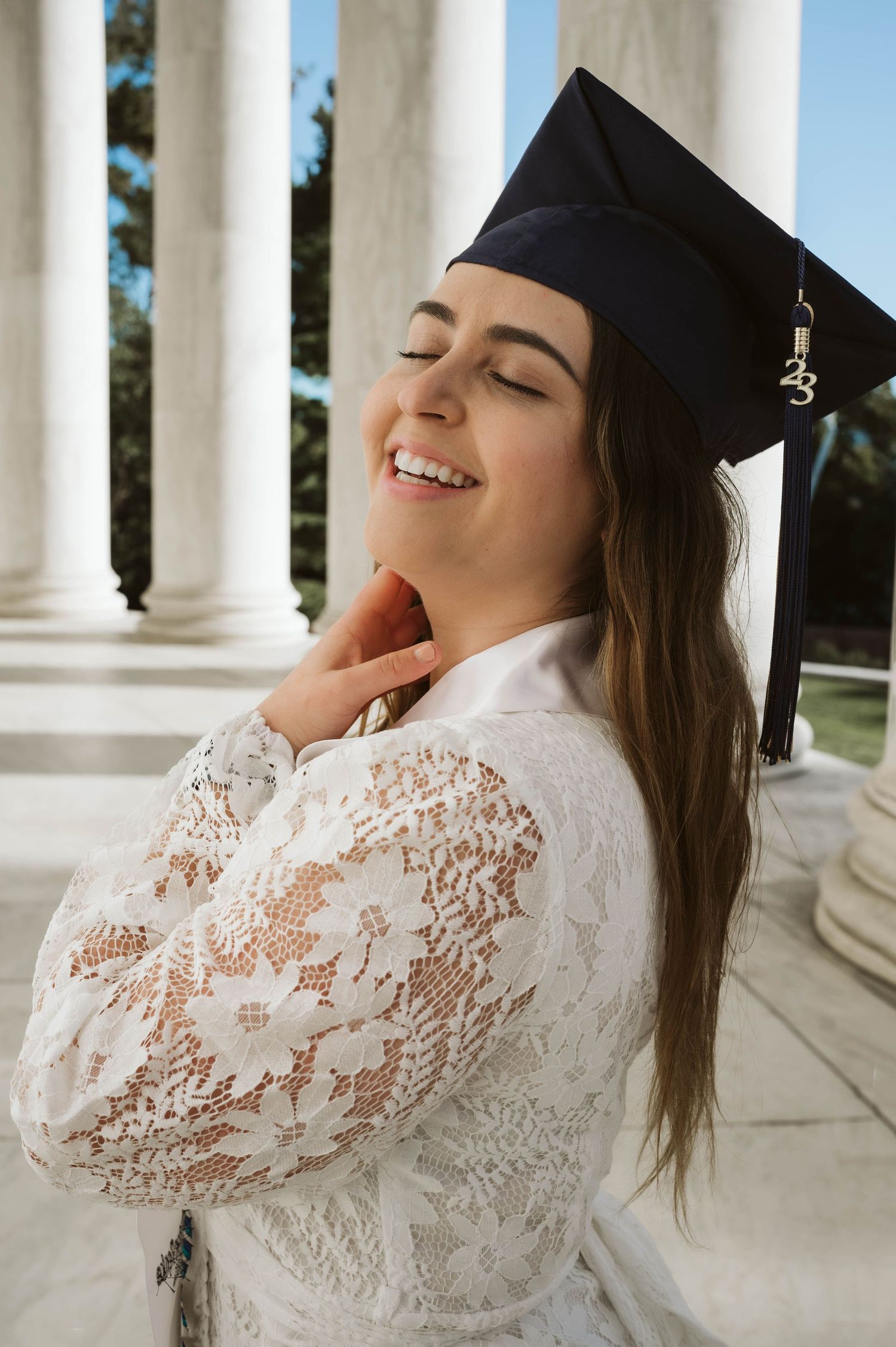 A graduate in cap and white lace dress smiles joyfully while posing near classical columns on graduation day.