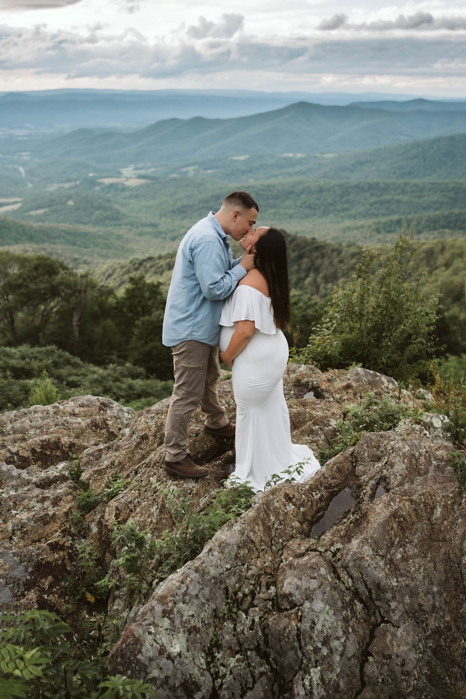A couple shares a romantic moment atop a mountain peak overlooking a misty valley of rolling hills and forests.