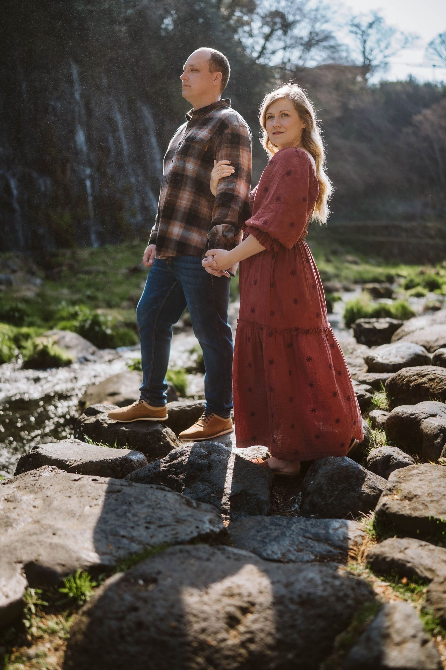 Two children stand together on rocky terrain near a stream while sunlight filters through trees in the background.