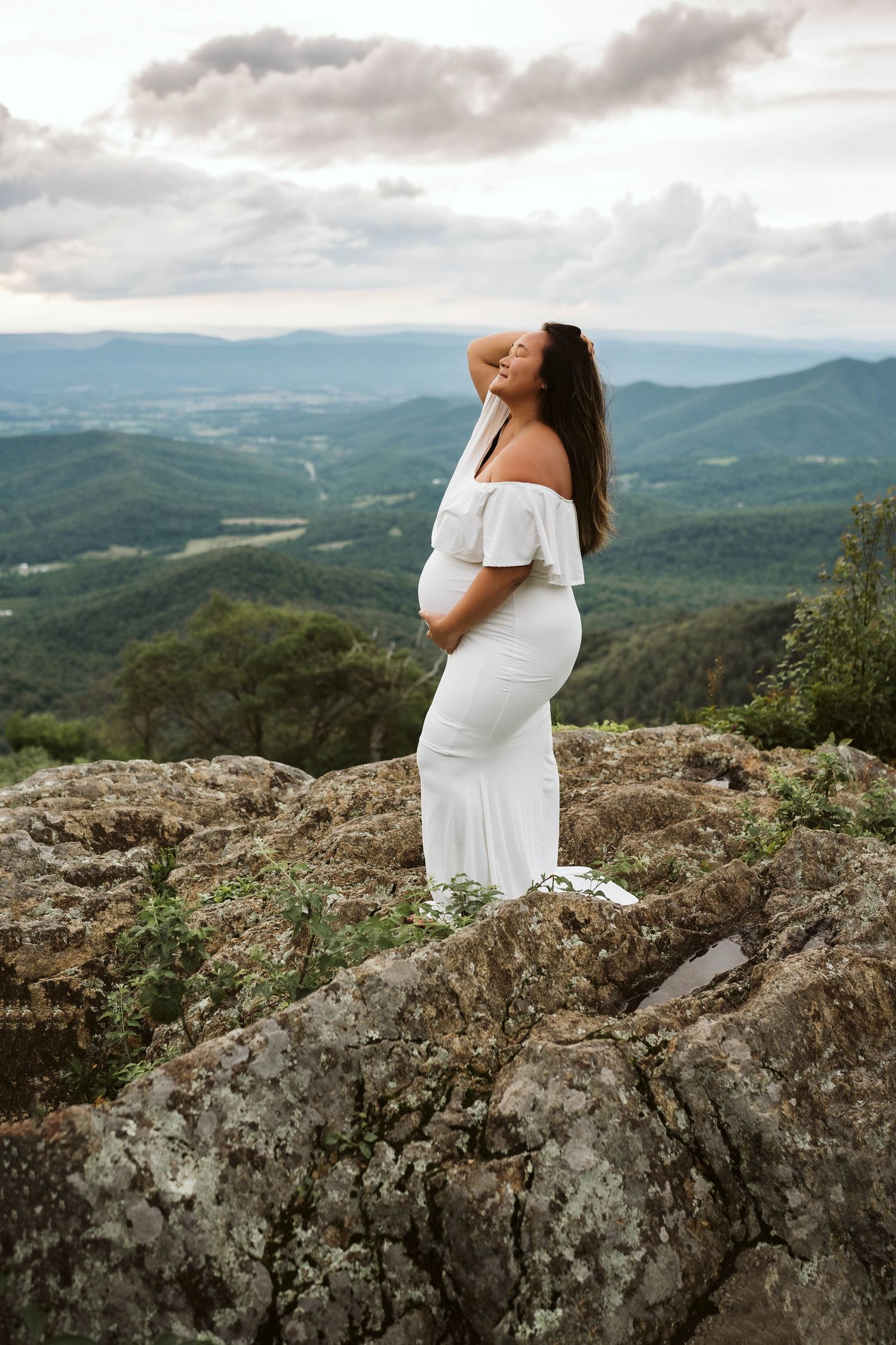 Person in flowing white dress stands on rocky mountain overlook with misty blue ridges and valley stretching into distance.