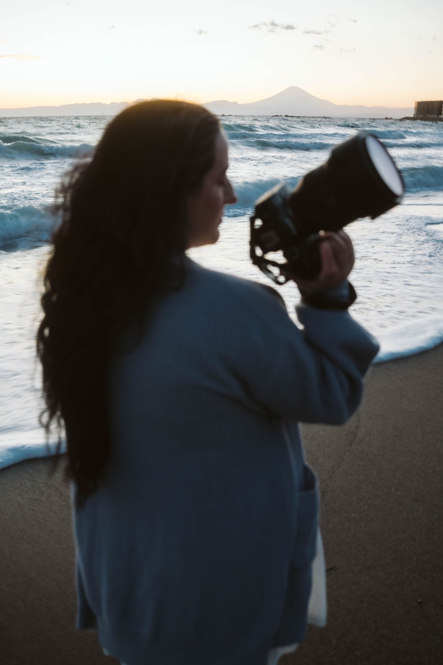 A silhouette holding a camera on a beach at sunset with waves and mountains in the distance.