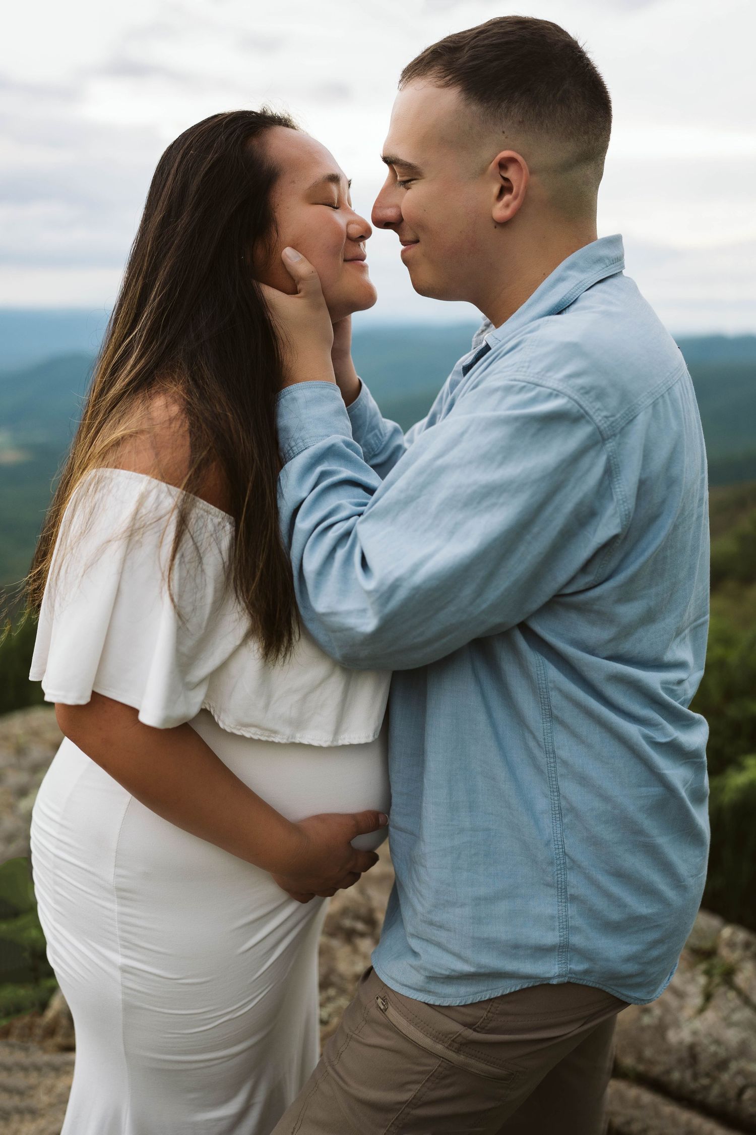 A couple shares an intimate moment on a mountain overlook, with the woman wearing white and the man in light blue.