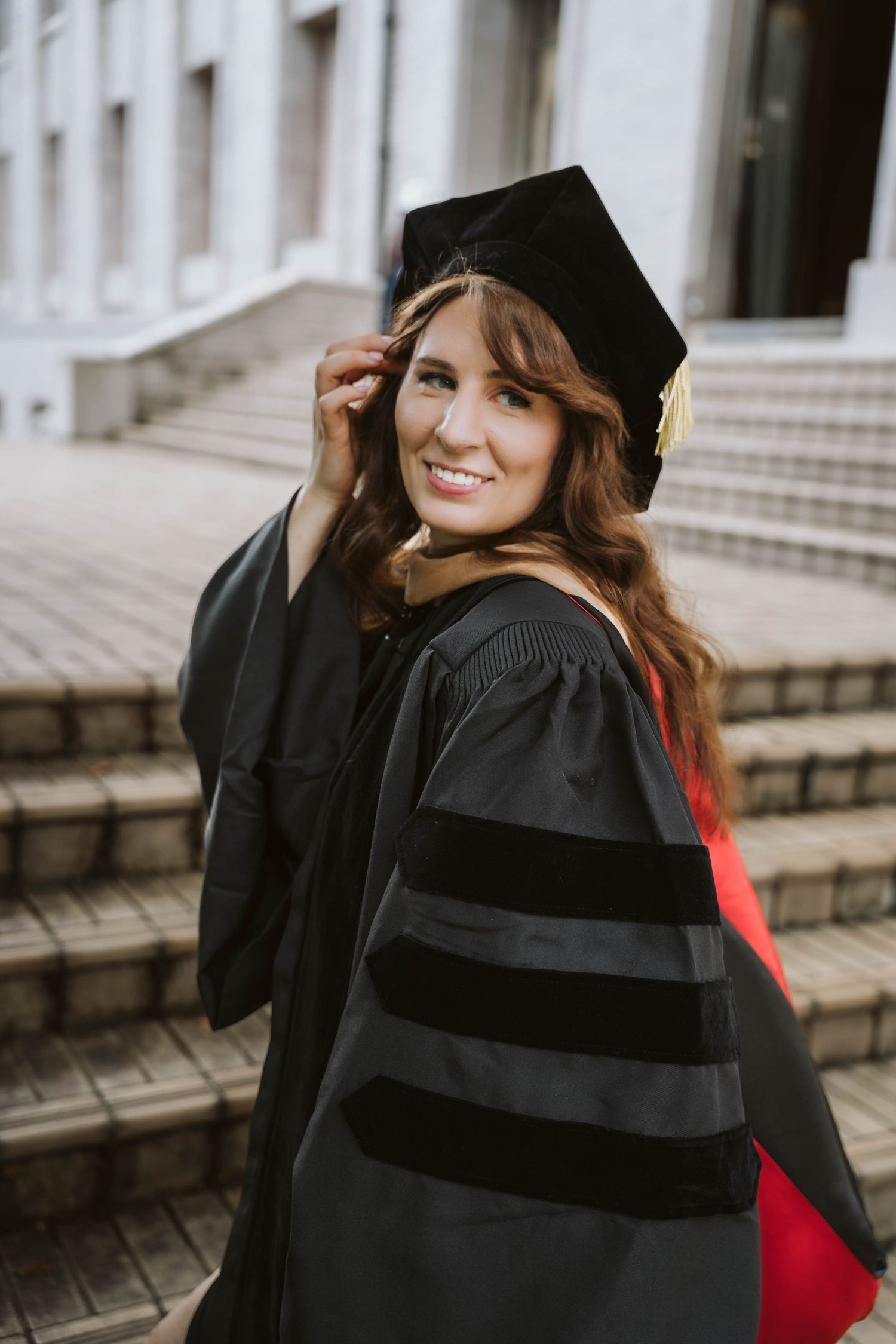 Graduate in black cap and gown with red stole smiles during outdoor graduation photoshoot.