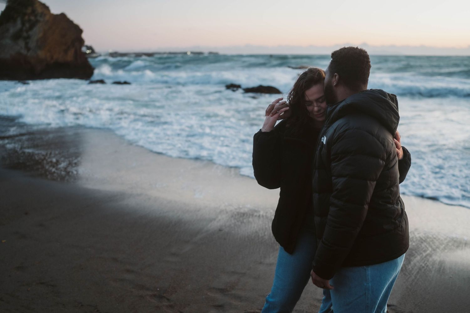 Two silhouettes embrace on a dark beach at sunset with crashing ocean waves in the background.