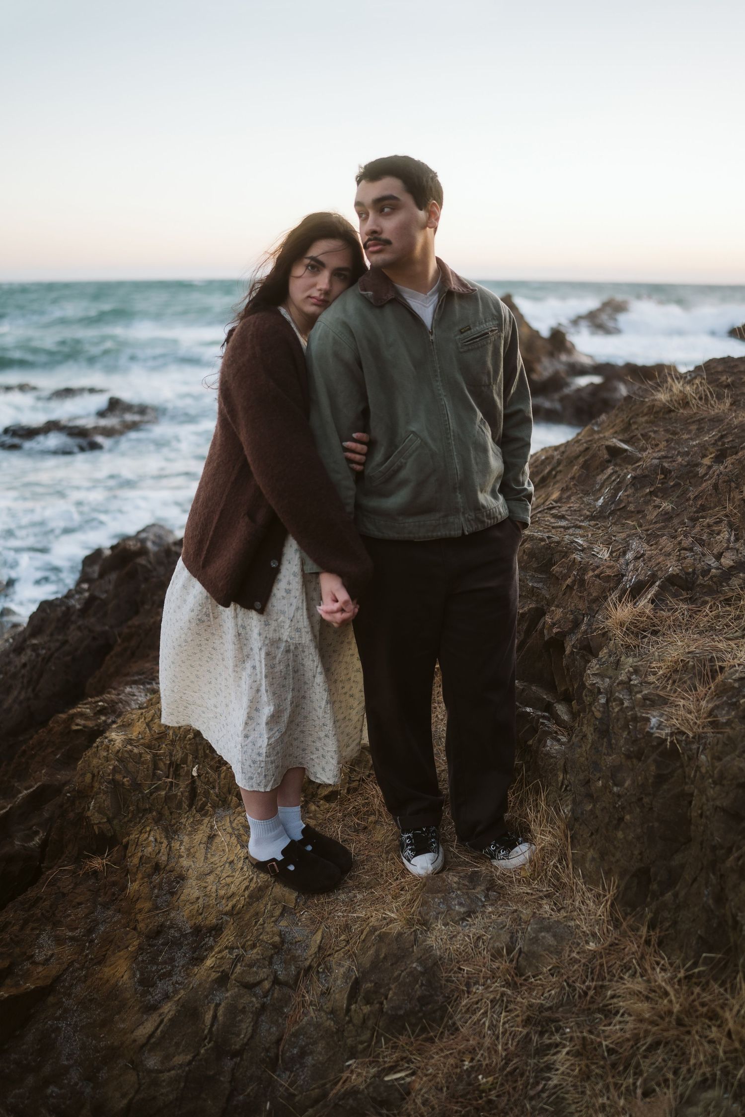 A couple shares an intimate moment on rocky coastal cliffs at sunset with crashing waves in the background.