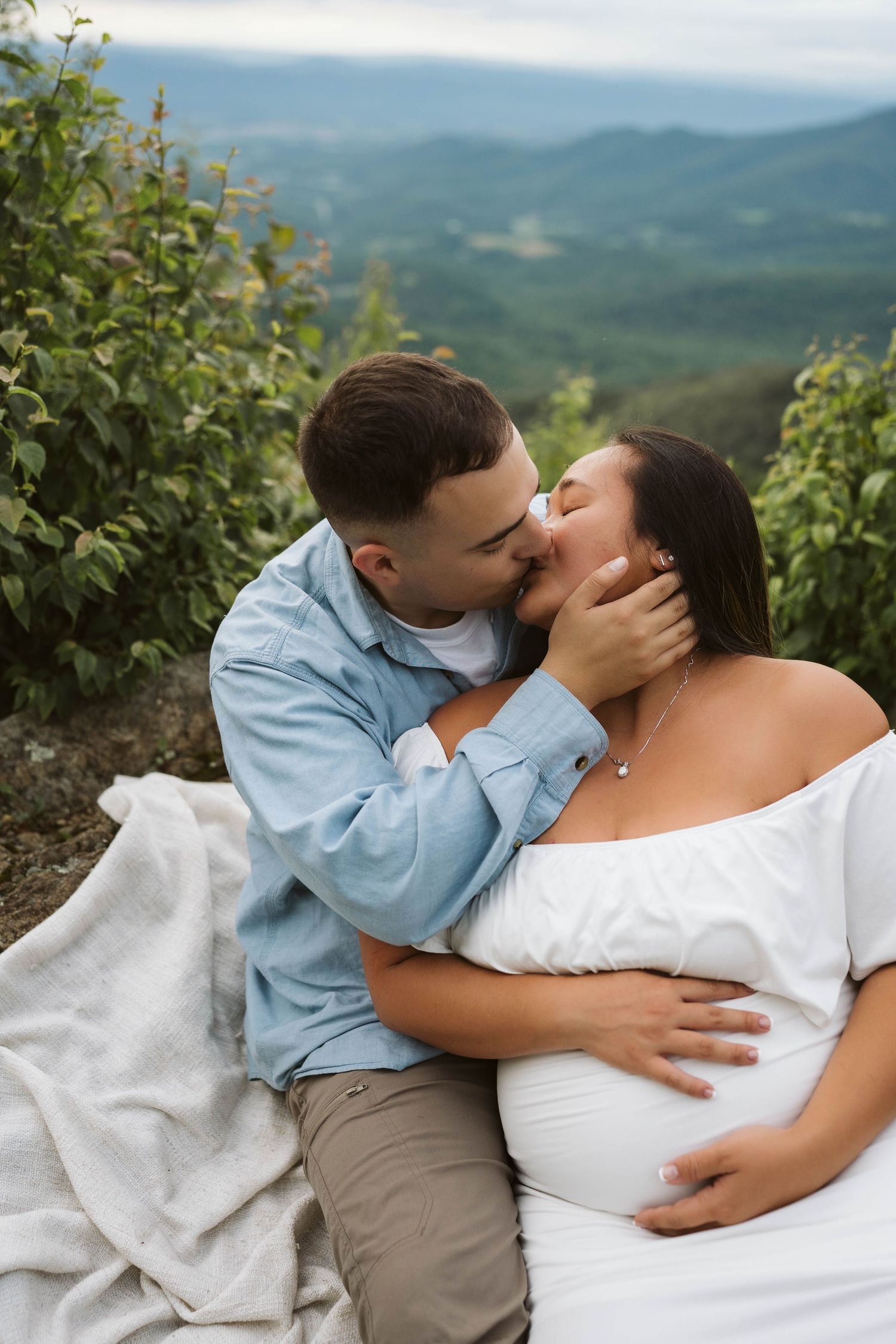 Couple shares intimate moment on mountaintop with scenic green mountain landscape in background.