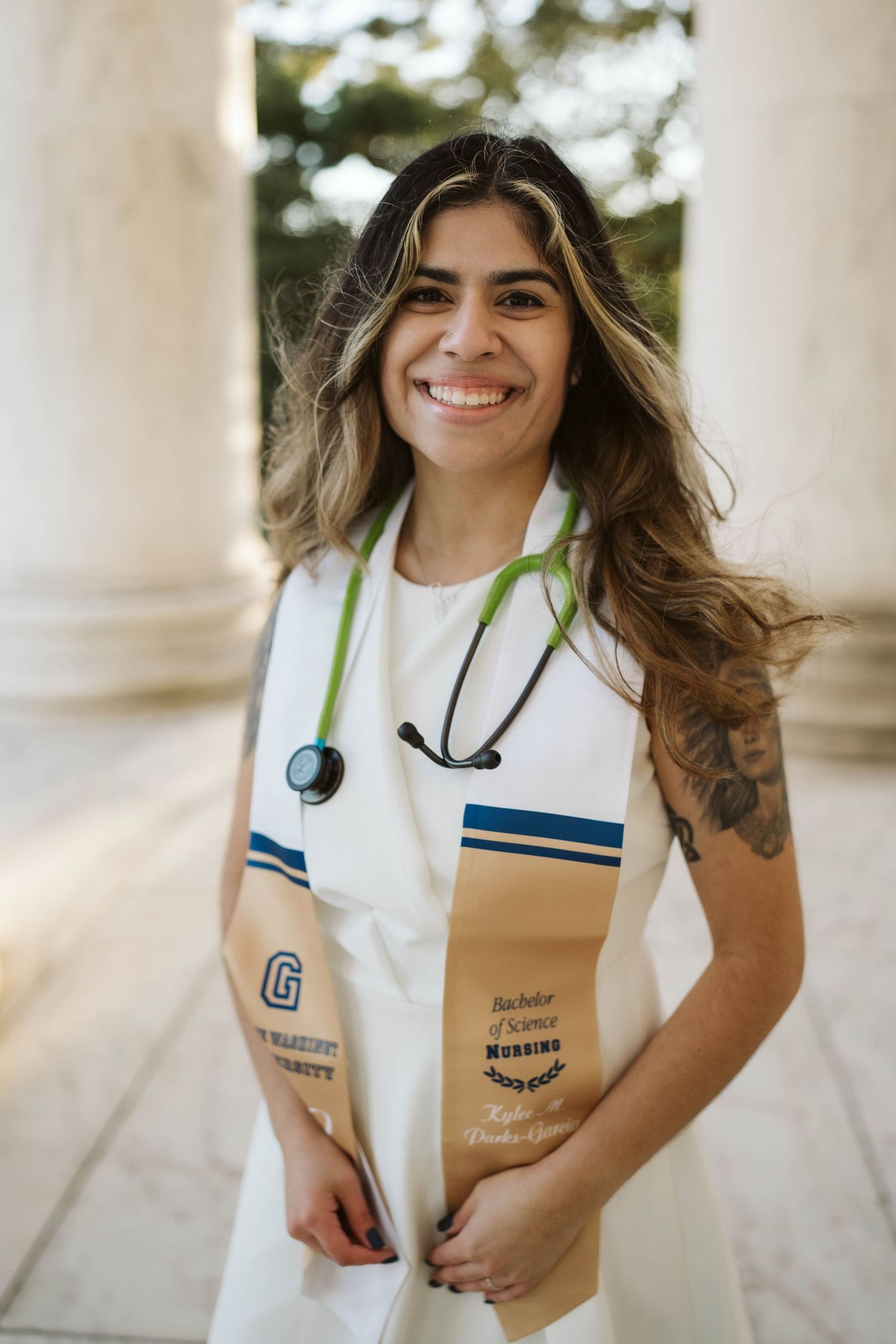 Healthcare graduate wearing stethoscope and white graduation stole with blue stripes smiles during outdoor portrait session.