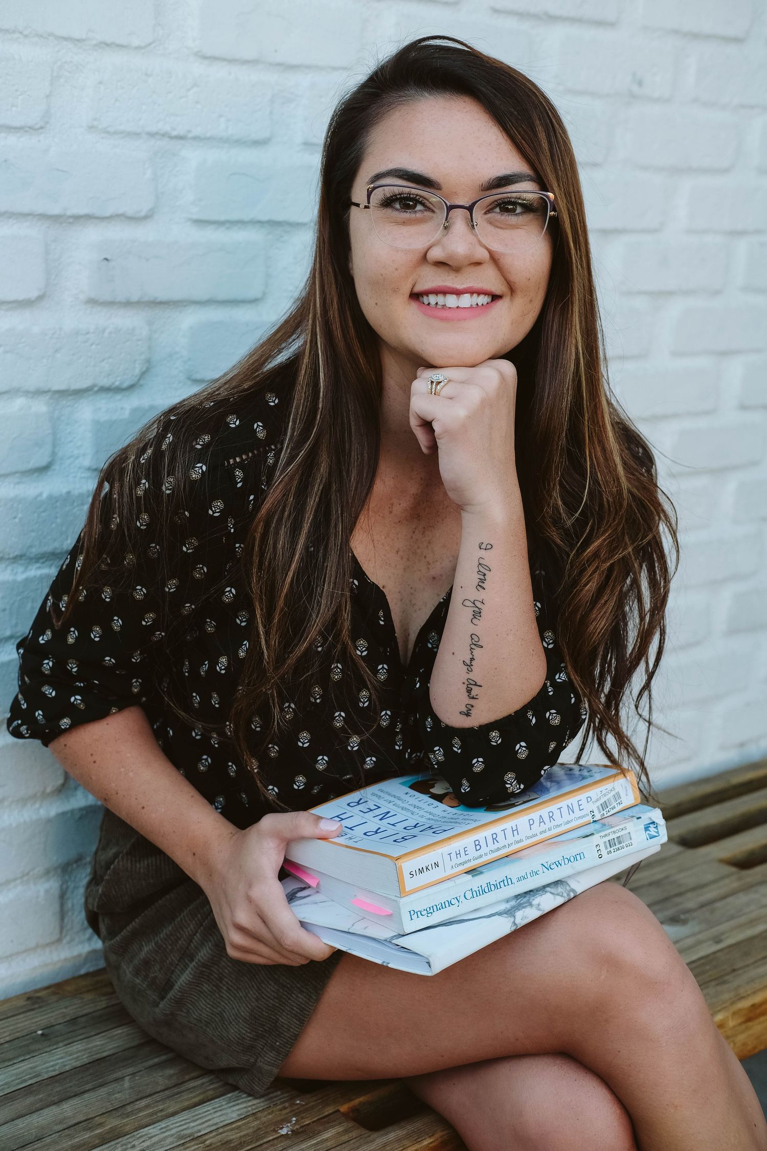 A person in glasses and a dark patterned top sits on a wooden bench holding books with a white brick wall background.