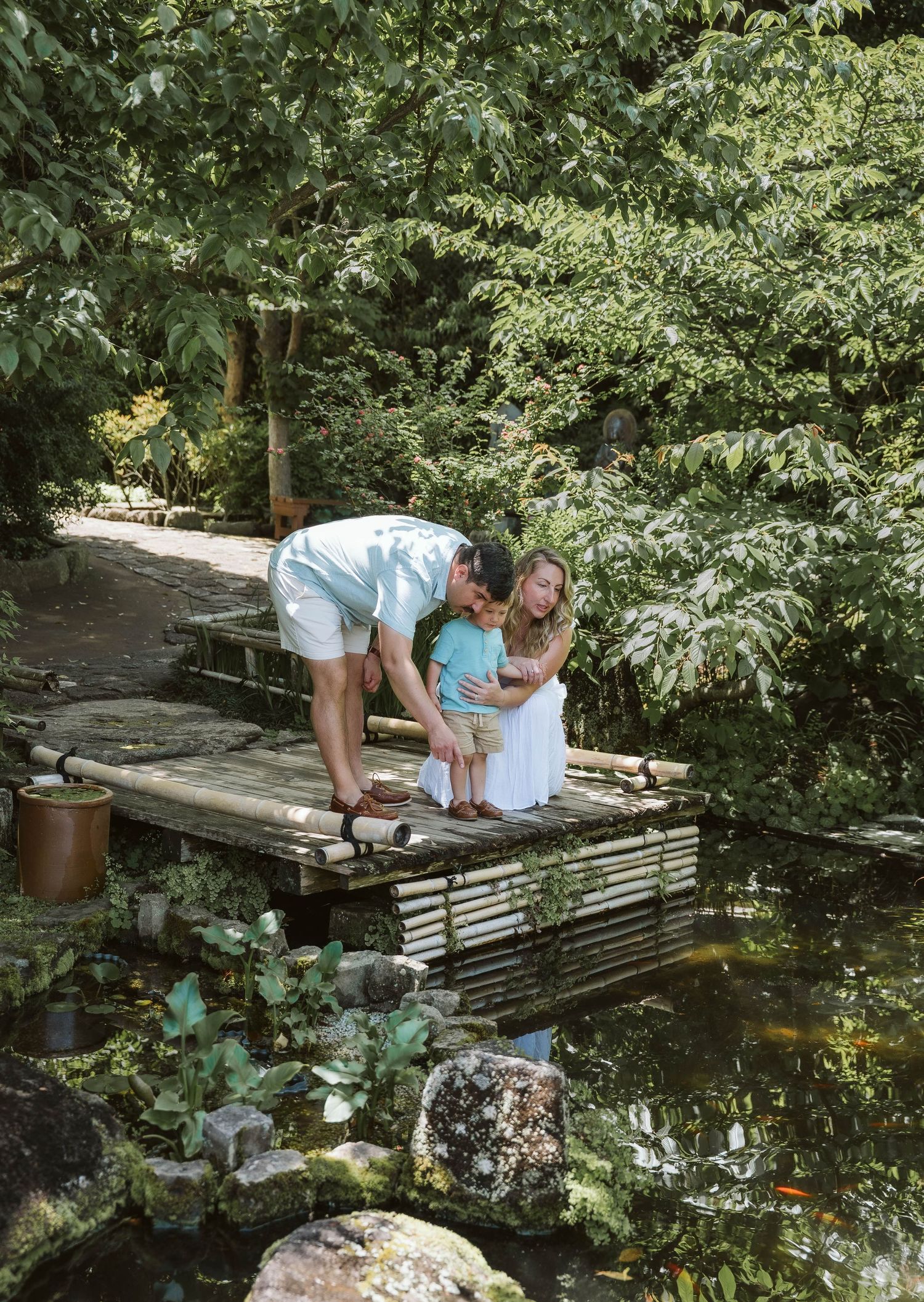 People leaning over a wooden deck to observe fish swimming in a tranquil garden pond surrounded by lush greenery.