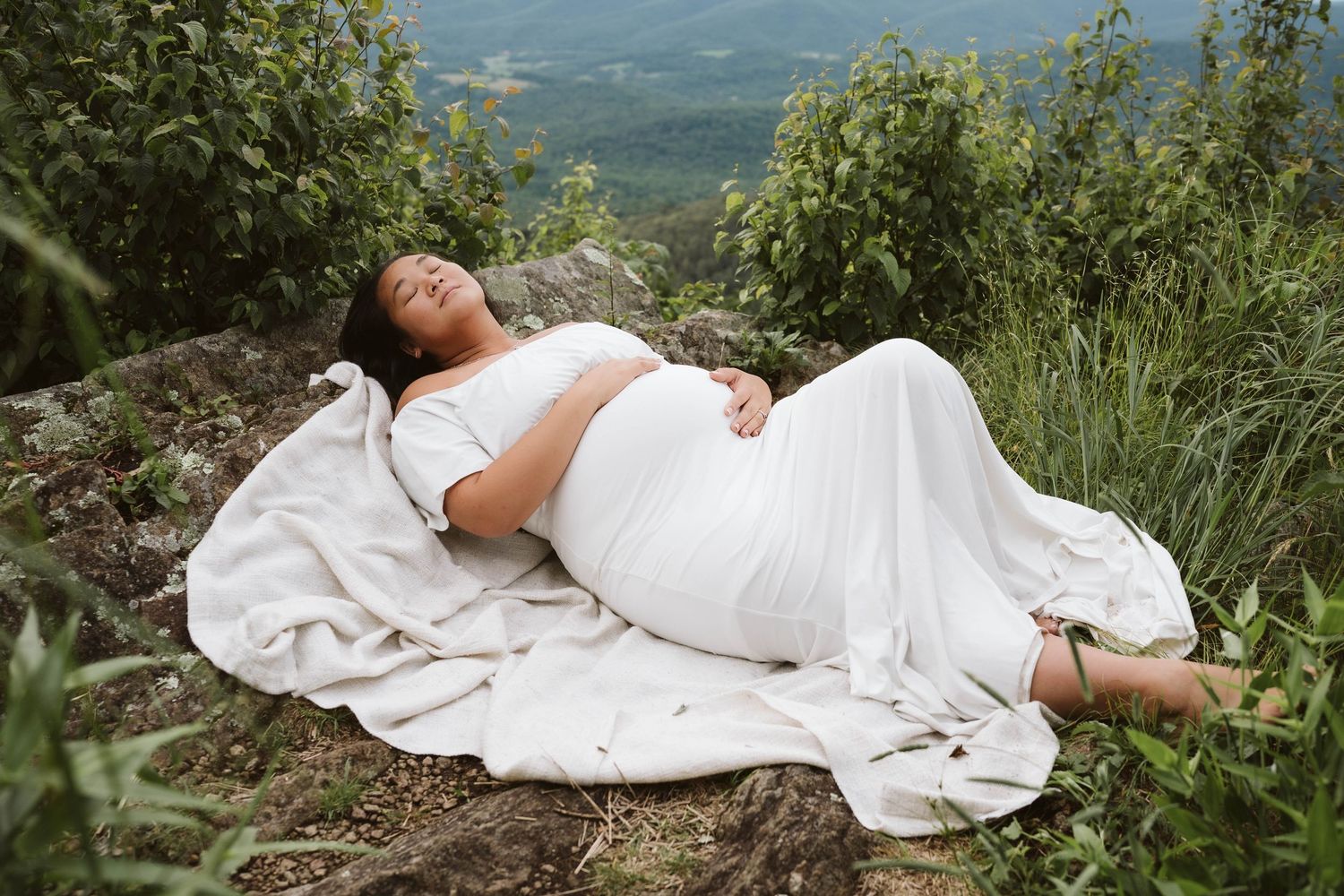 Person in flowing white dress lying peacefully on blanket atop mountain overlook surrounded by greenery.