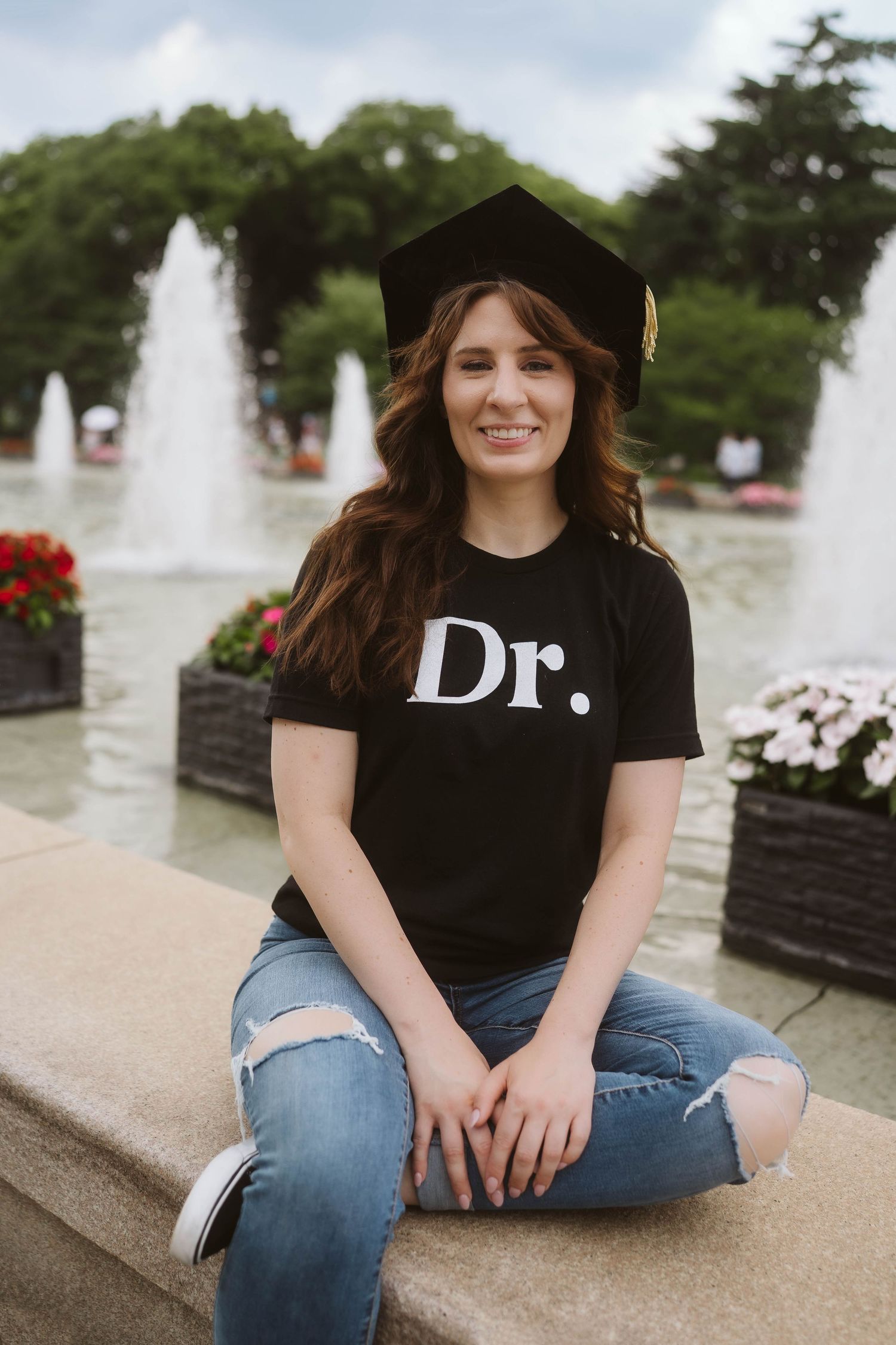 Graduate wearing academic cap sits on stone ledge near fountain with flowers in background.