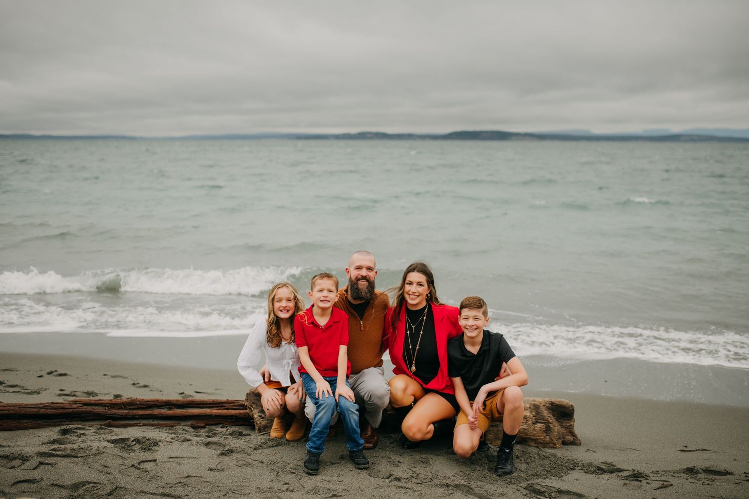 Family of five smiling together on a beach with ocean and cloudy sky behind them.
