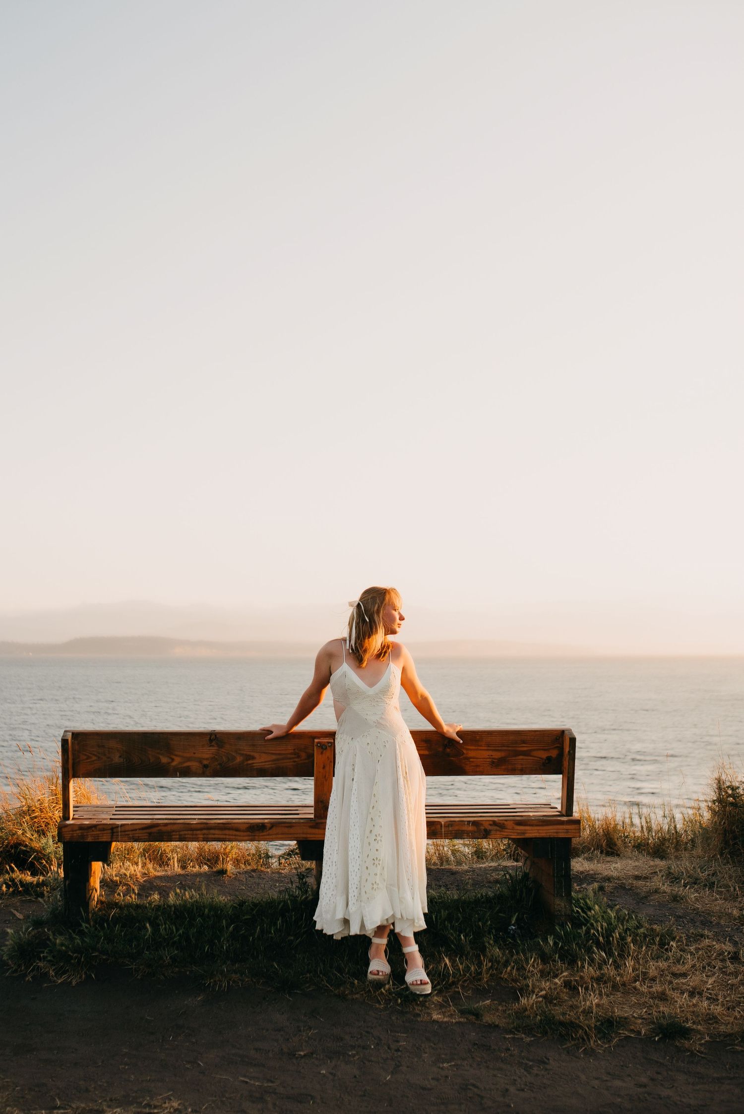Blonde woman in white lace dress standing at a wooden bench overlooking the ocean at sunset.