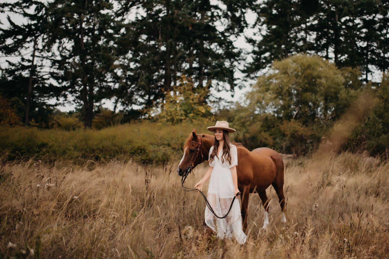 Young woman in white dress and sun hat walking with chestnut horse through dry grass field.
