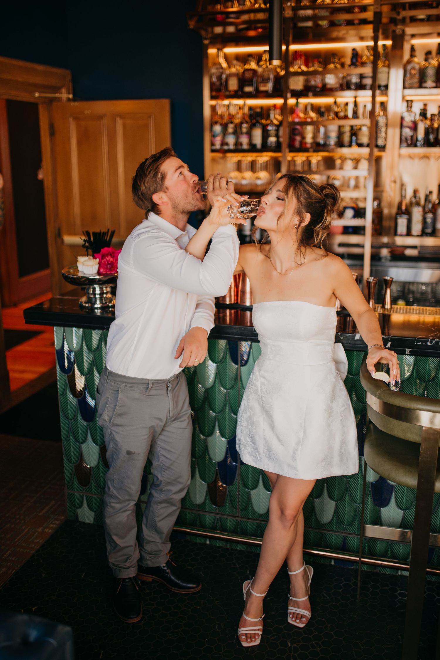 Couple laughing and drinking at a stylish bar with green tiled counter and backlit bottle display.