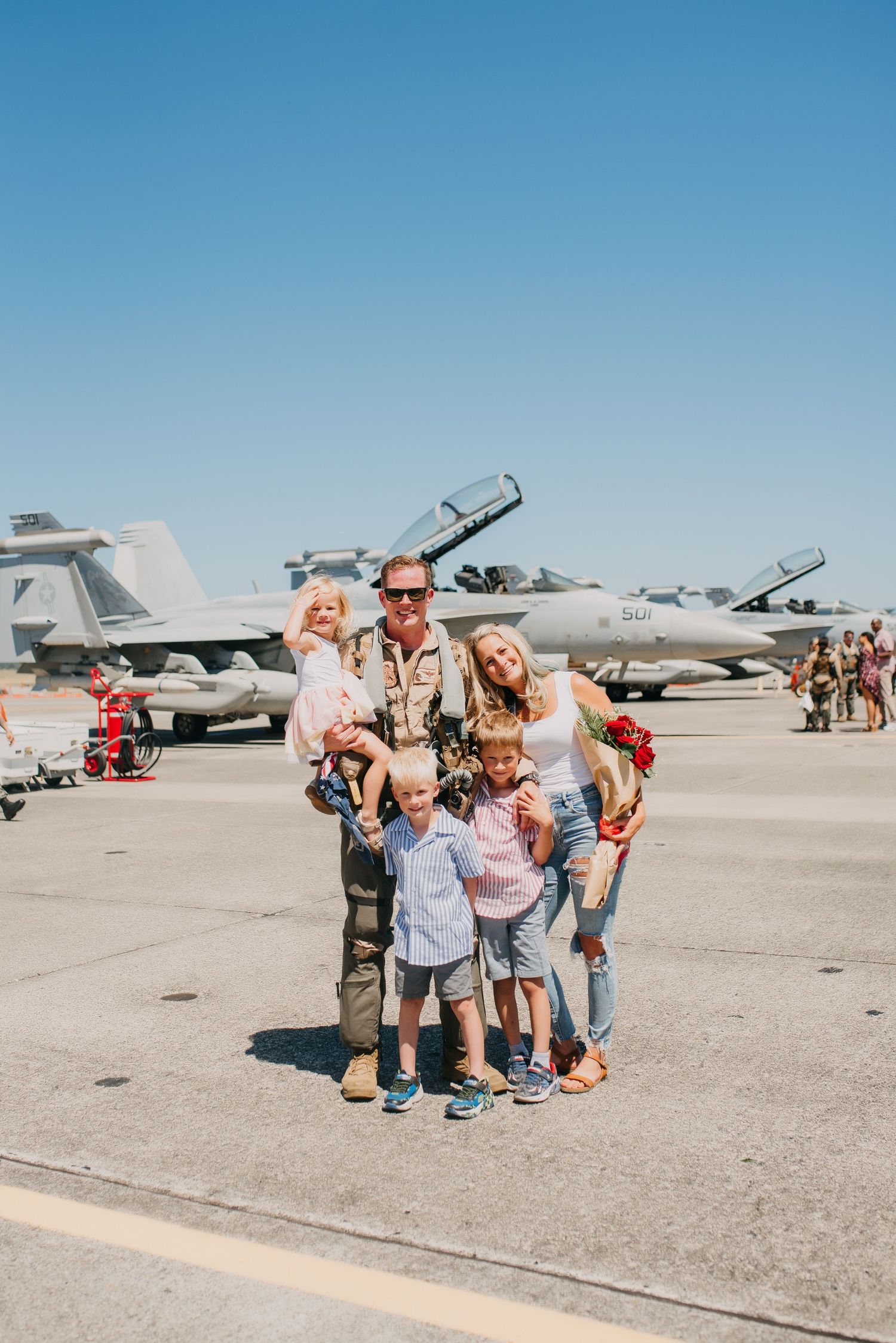 Military family reunion on airfield tarmac with fighter jets, father in flight suit holding children with wife beside him.