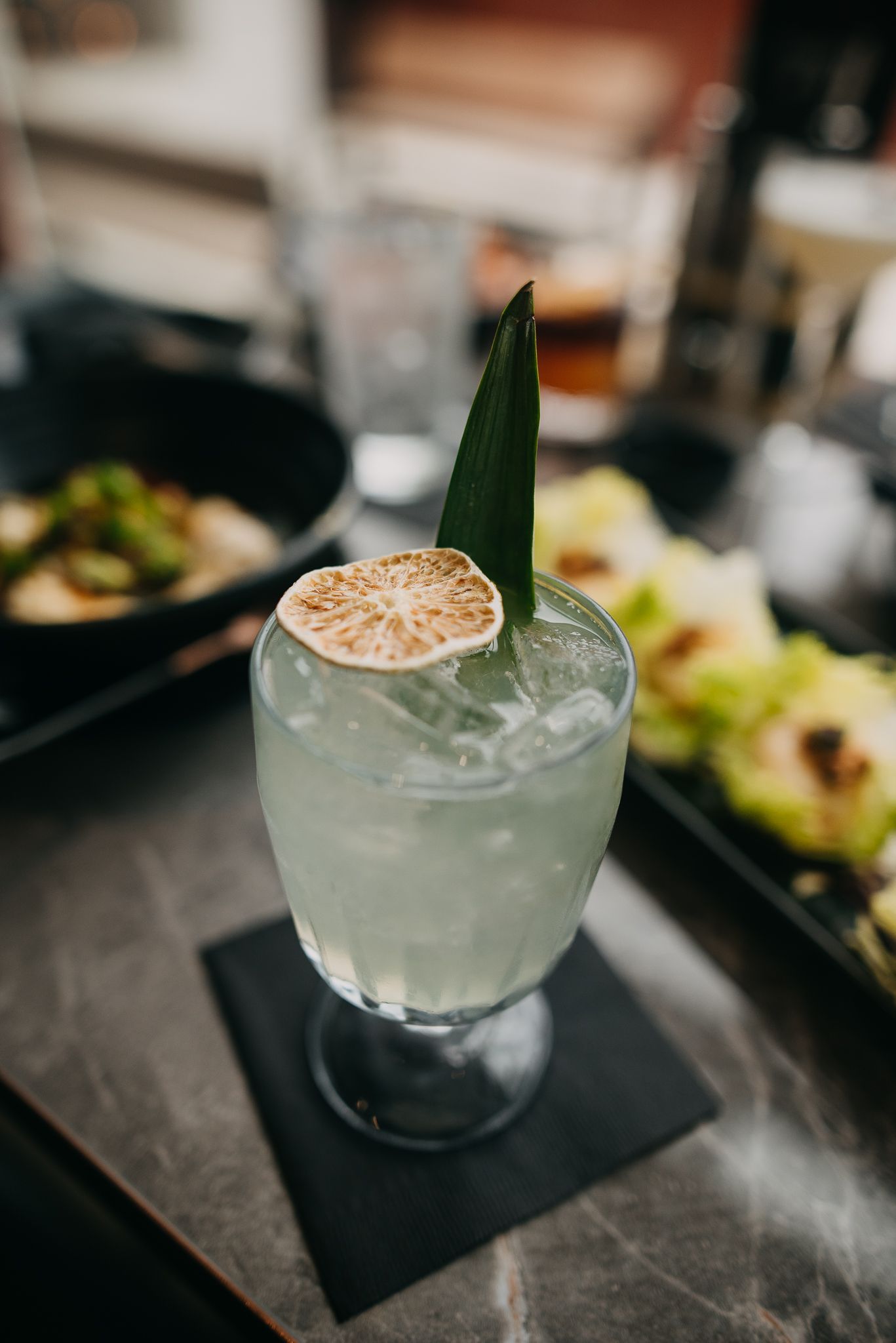 Refreshing cocktail with a dehydrated lime slice and pandan leaf garnish on a marble surface.