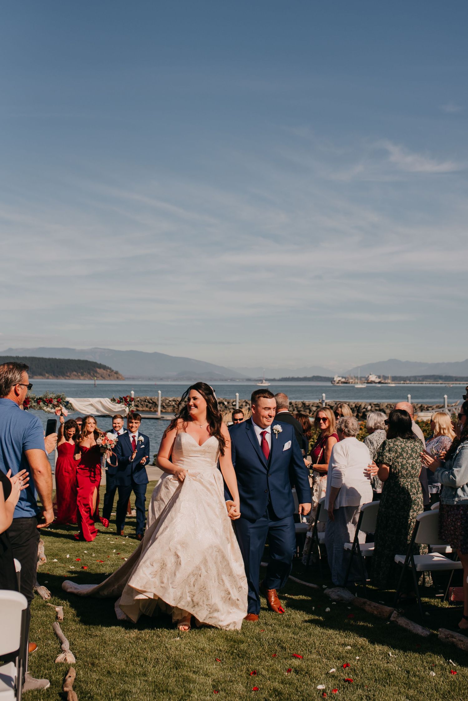 Newlyweds walk down the aisle at an outdoor waterfront wedding ceremony on a sunny day.