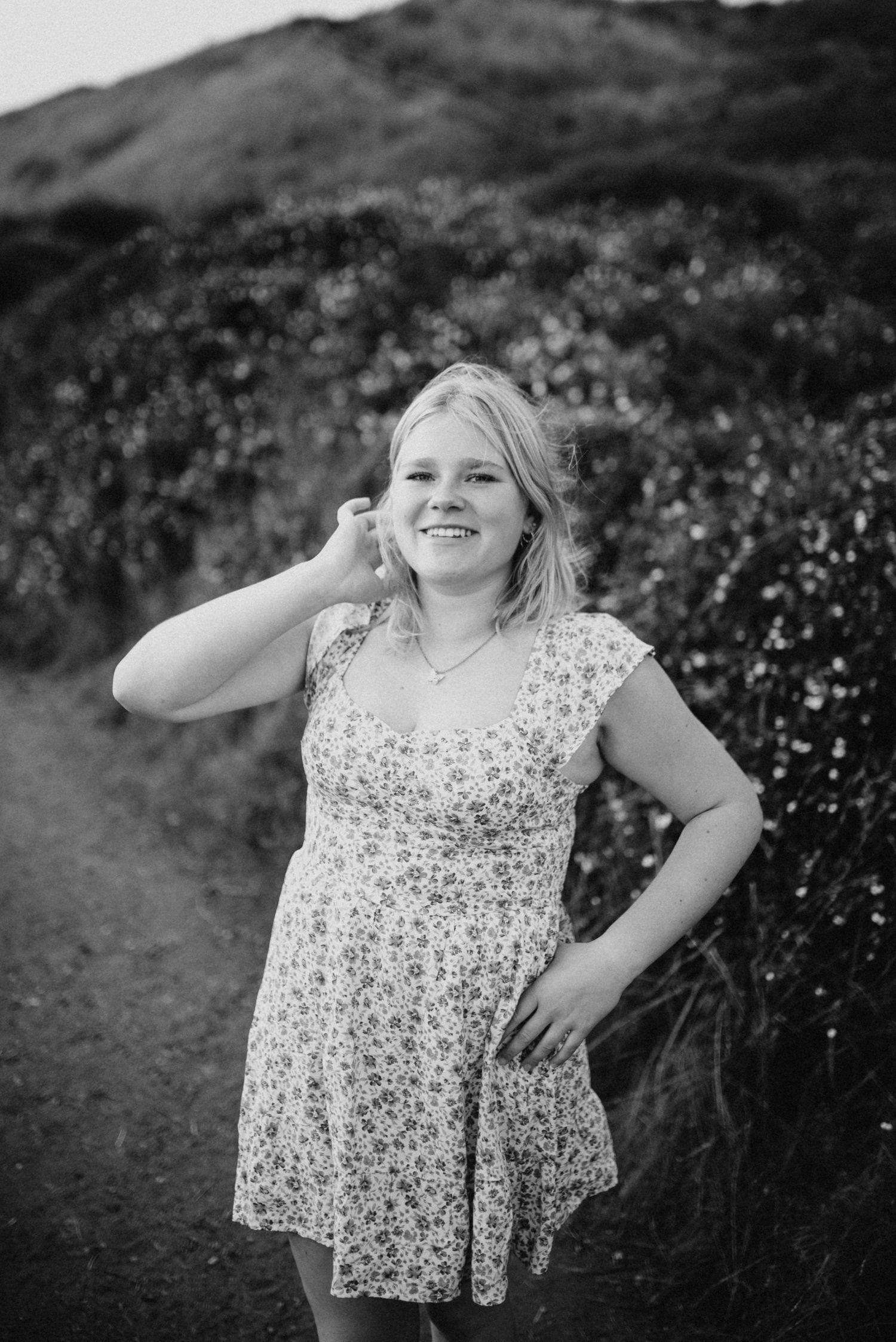 Smiling blonde teen girl in floral dress posing on nature trail in black and white senior photo.