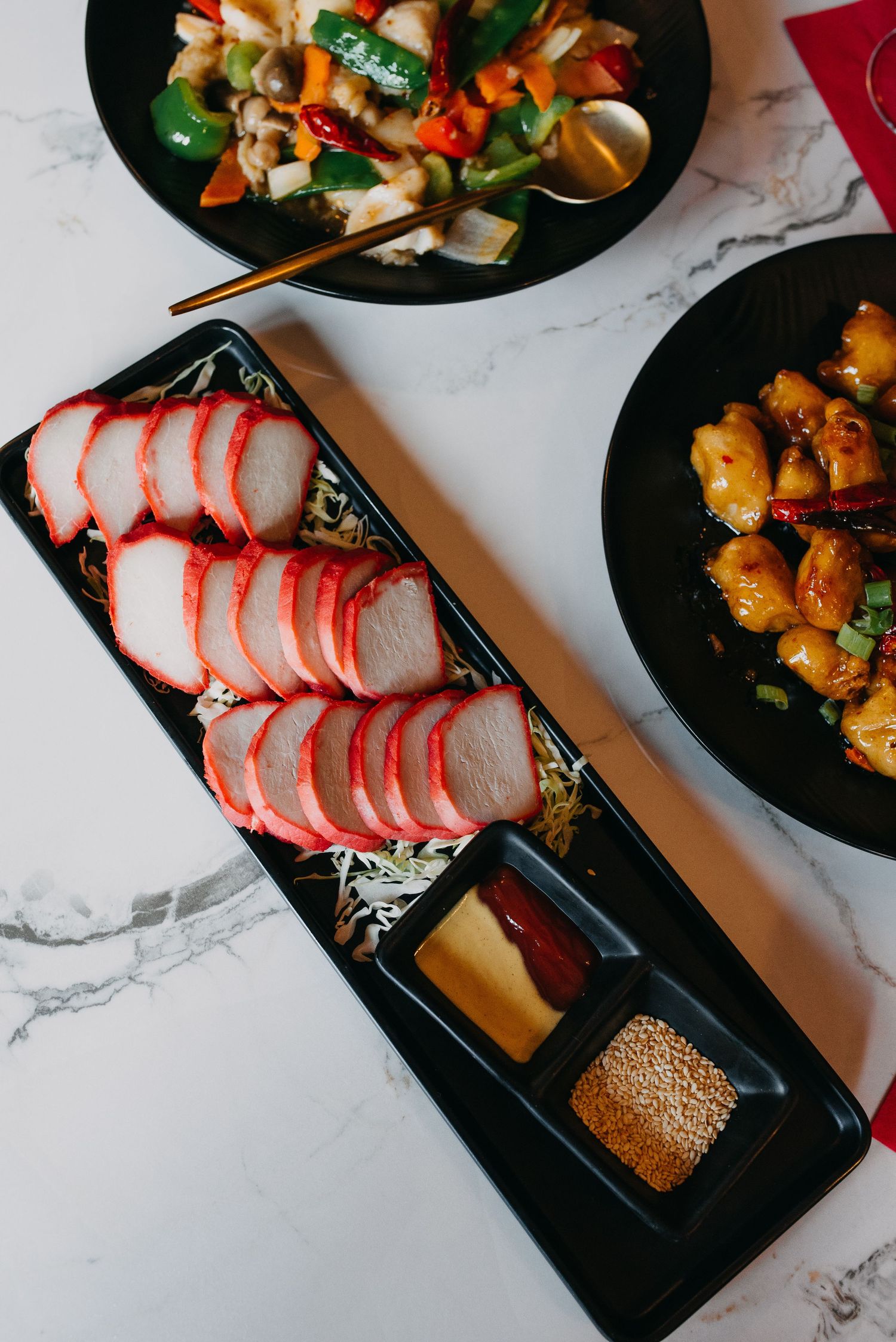 Sliced char siu pork with dipping sauces on a black plate alongside stir-fried dishes on marble table.