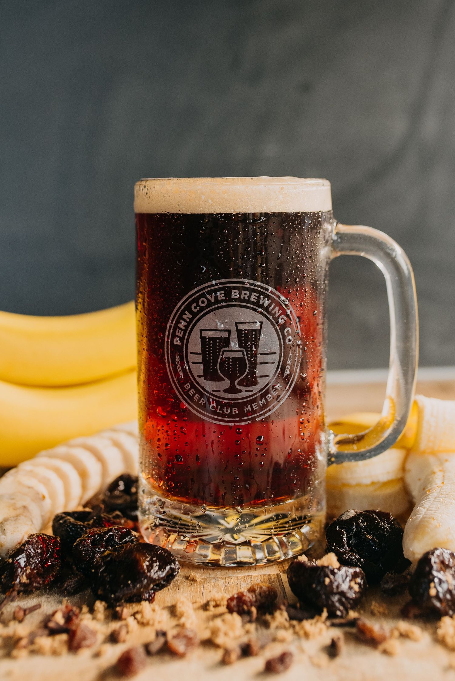 Dark craft beer with frothy head in a branded Finn Cove Brewing Co. mug surrounded by bananas and dried fruit.